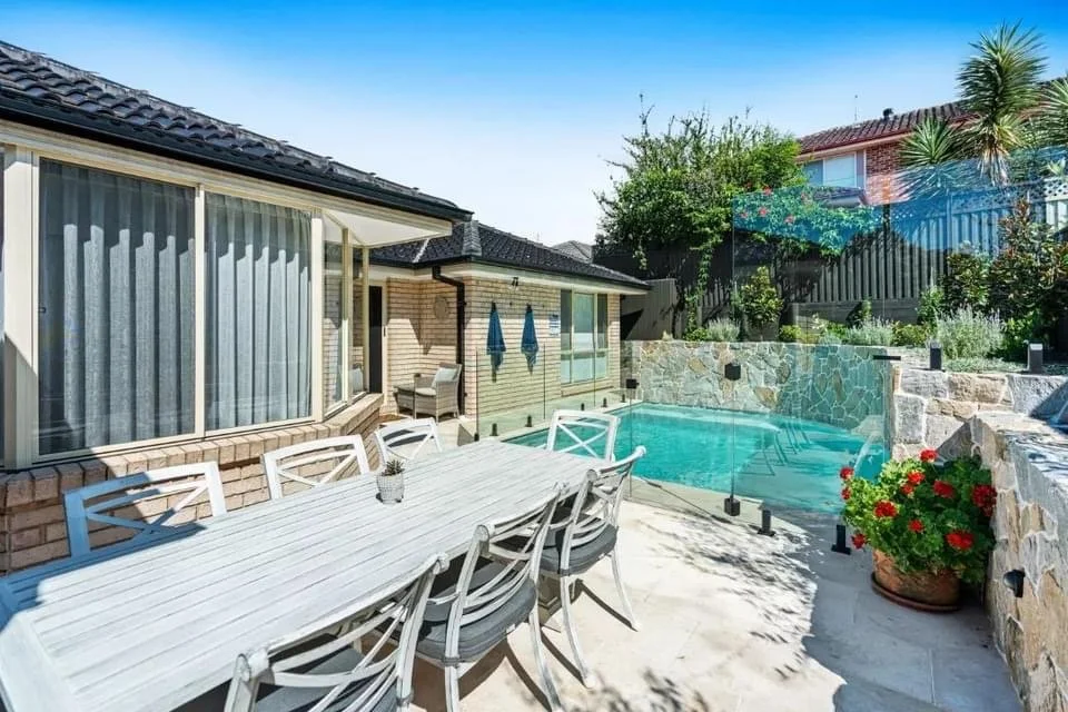 Backyard with a swimming pool, patio dining area with a large white table and chairs, house with sliding glass door, lush greenery, and a stone wall.
