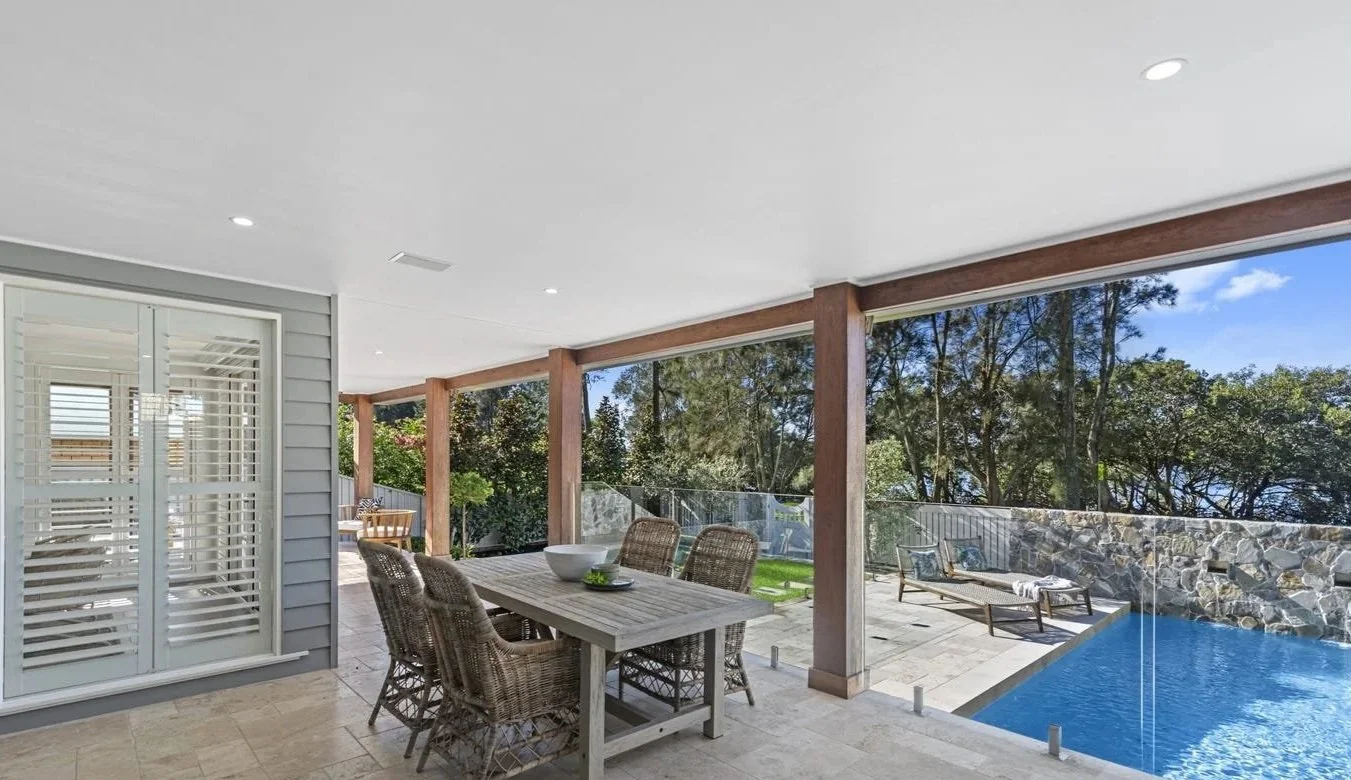 Covered outdoor patio with dining table, wicker chairs, poolside loungers, and a swimming pool, with trees and sky in the background.