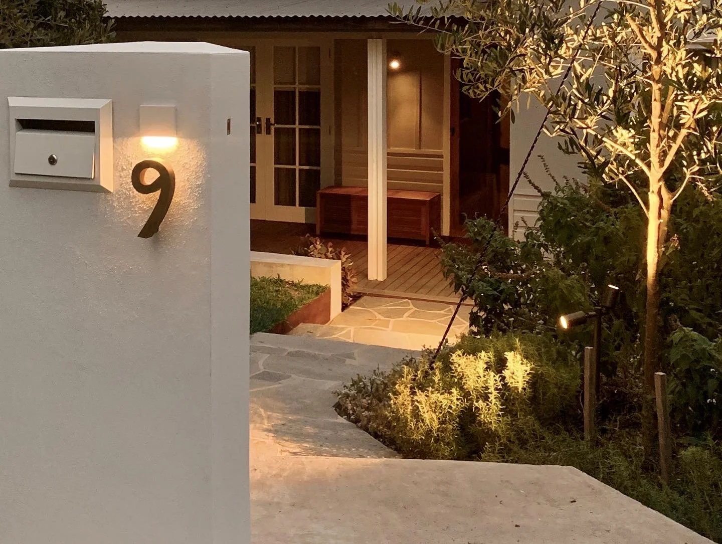 Modern house with a white wall featuring a black number 9, a mailbox, and a light. The front door is made of glass and wood, leading to a porch with a bench, surrounded by greenery and pathway lights at dusk.