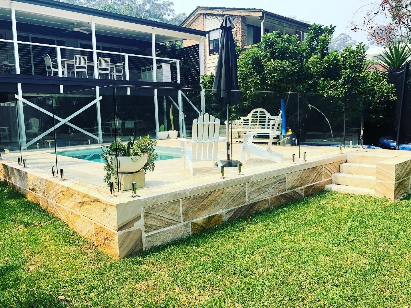A backyard with a small pool enclosed by a glass fence, surrounded by white outdoor furniture and potted plants, with a house in the background and a grassy lawn in the foreground.