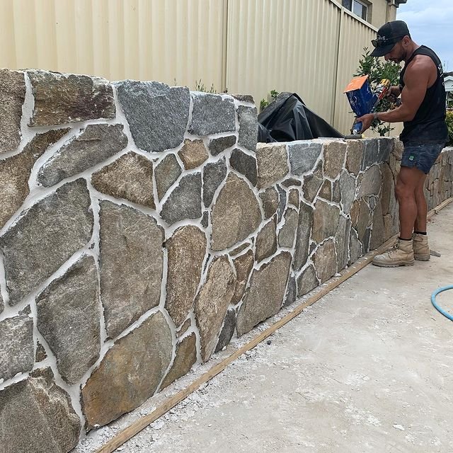 A man wearing sunglasses, a sleeveless shirt, shorts, and work boots building a stone wall outside. He is holding a bag of mortar or grout and working on the stone wall, which is constructed with irregular stones and white mortar. There is a yellow f