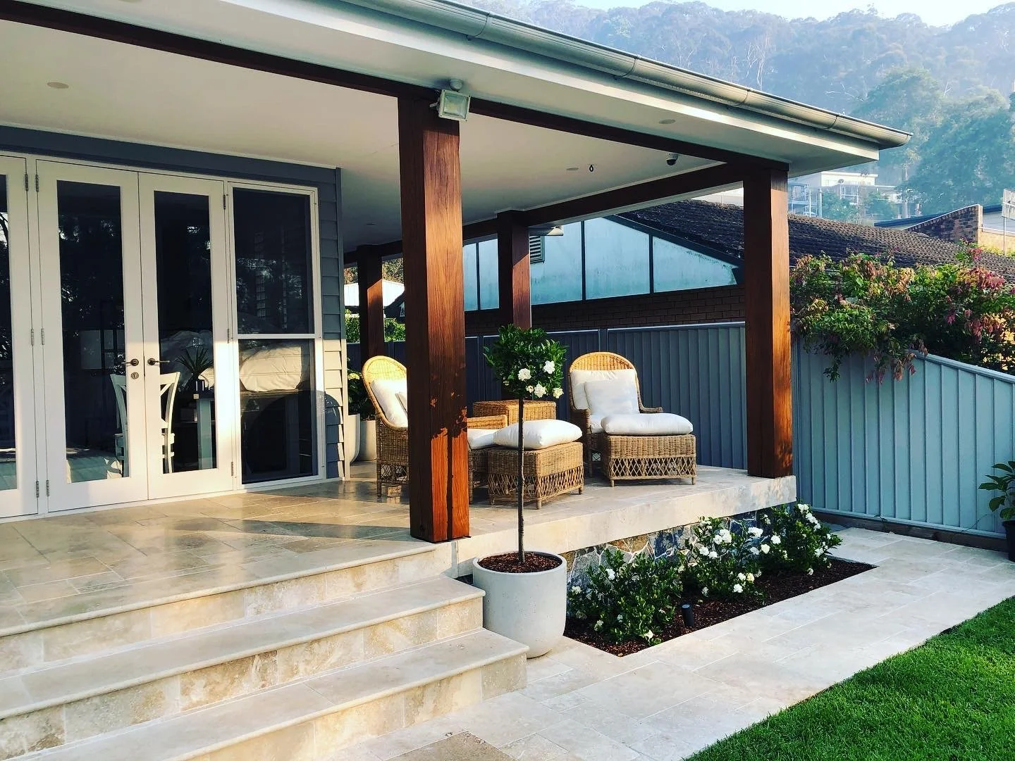 Back porch with wicker chairs, potted plants, sliding glass doors, and a flower garden, surrounded by a metal fence.
