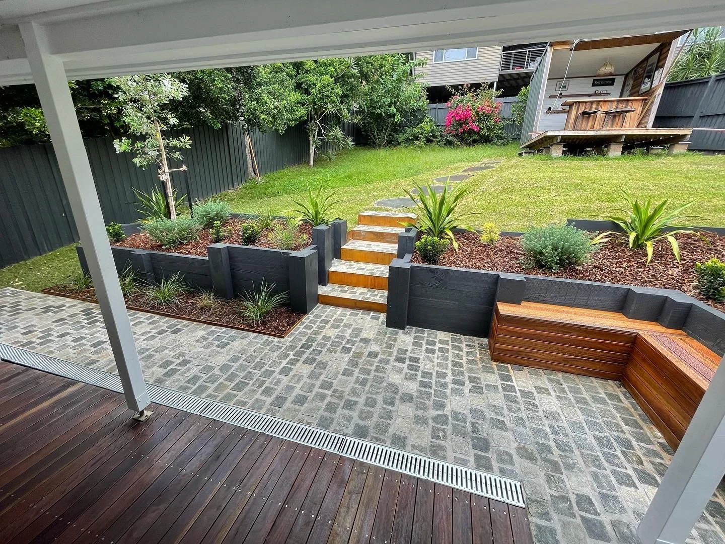 View of a backyard garden with a stone and wooden stairs leading to a grassy yard, surrounded by plants and trees, with a raised wooden deck in the background.