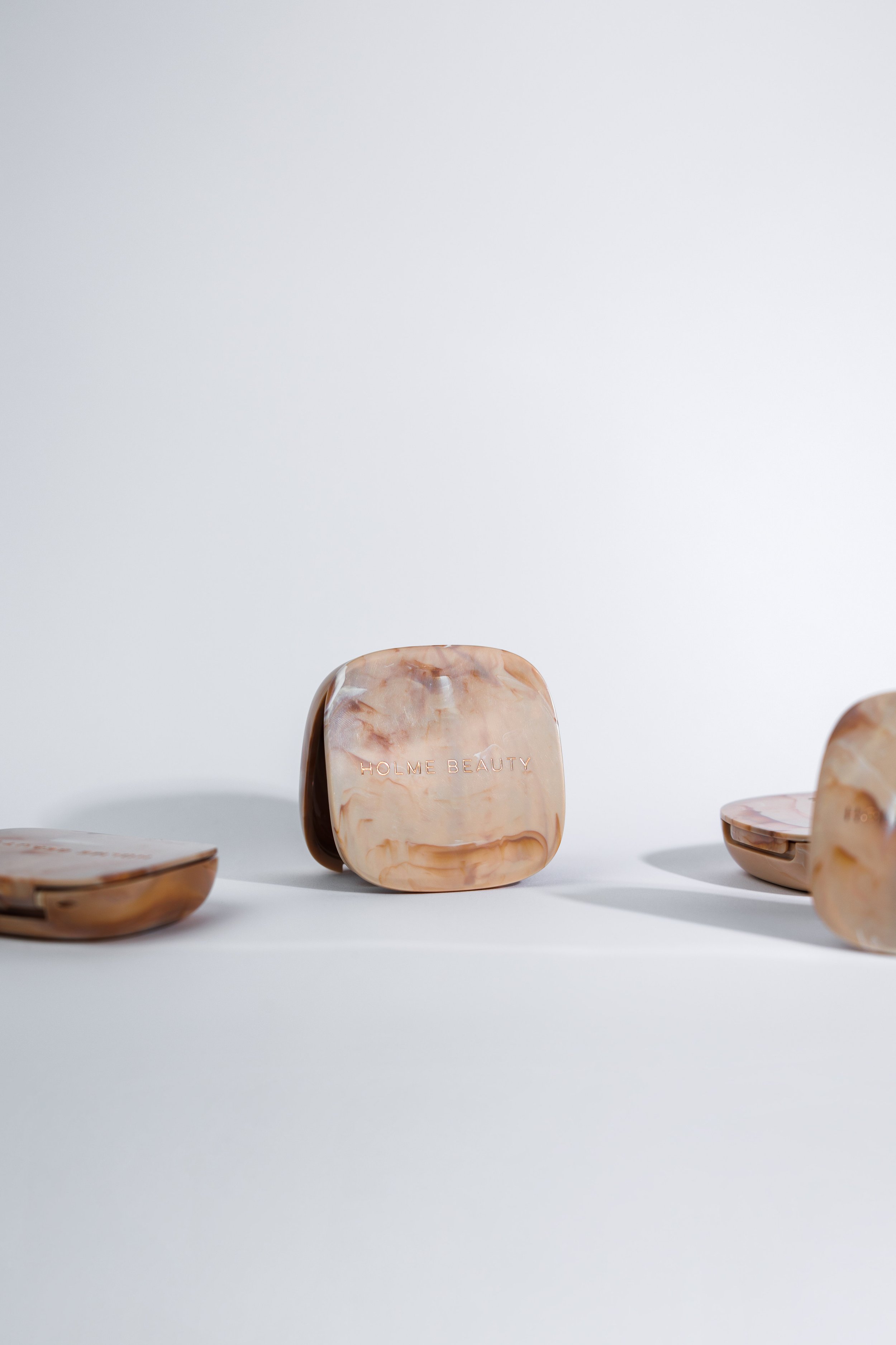 Three beige and brown marbled cosmetic containers on a white background, with the middle one displaying the text 'HOLME BEAUTY' engraved on it.