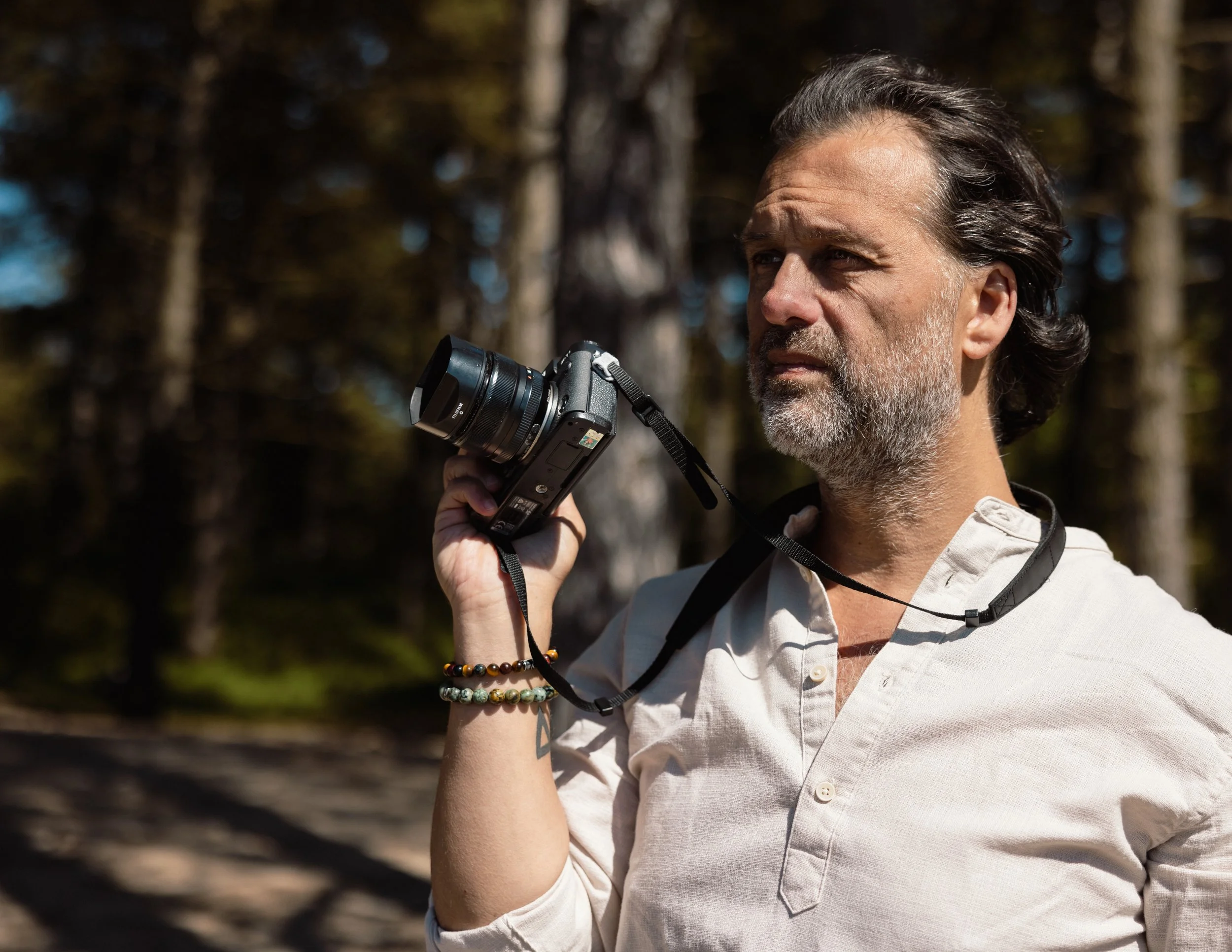 Man met lange, donker haar en een grijs gezichtshaar, gekleed in een witte blouse, houdt een digitale camera in de hand, buiten in een bosrijke omgeving met bomen op de achtergrond.