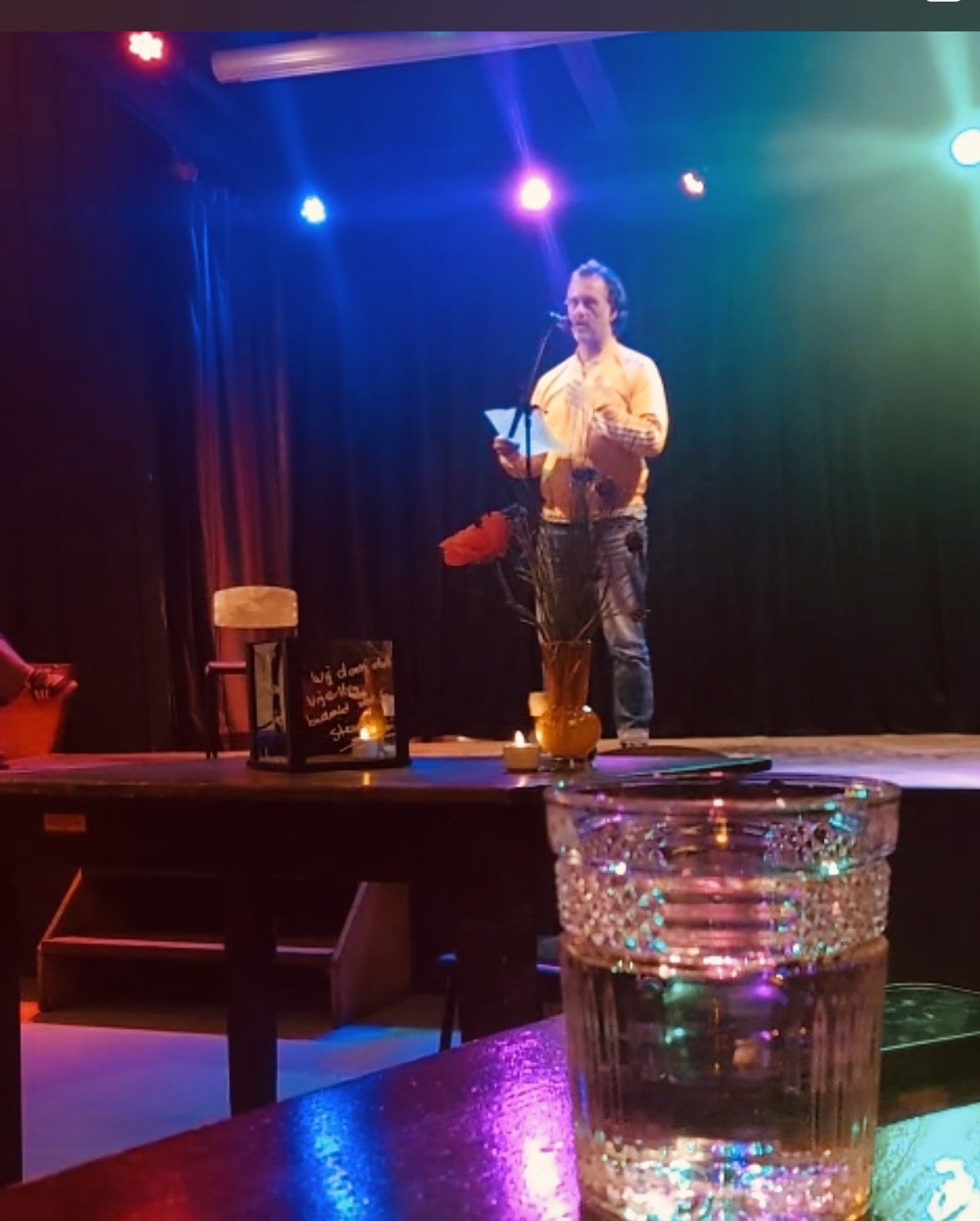 Man staat op een podium met een microfoon, omringd door gekleurde lichten, met een tafel en bloemen voor de podium en een glas water op de tafel in de voorgrond.