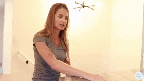 Kelly Sturm, a physical therapist, Wearing a gray shirt wipes a countertop with a cloth in a well-lit room, her careful movements resembling a breast cancer table stretch, reaching across the table to stretch her shoulder
