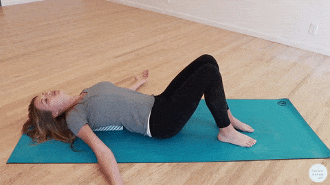 Kelly Sturm, a physical therapist, lies on her back on a yoga mat with knees bent and arms at her sides, a blue foam roller is under her, along her spine—a calming stretch often used in breast cancer recovery—amidst a bright, empty room with wooden floors.