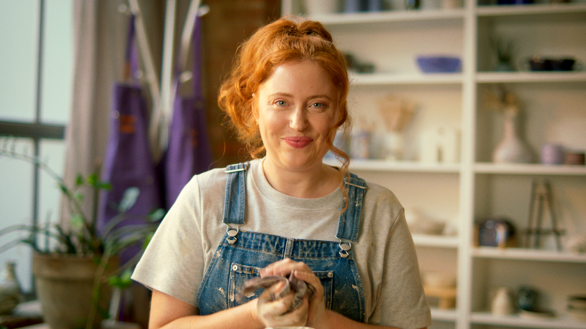 A young woman with curly red hair smiling and holding clay in a pottery studio for Multi-Gyn commercial
