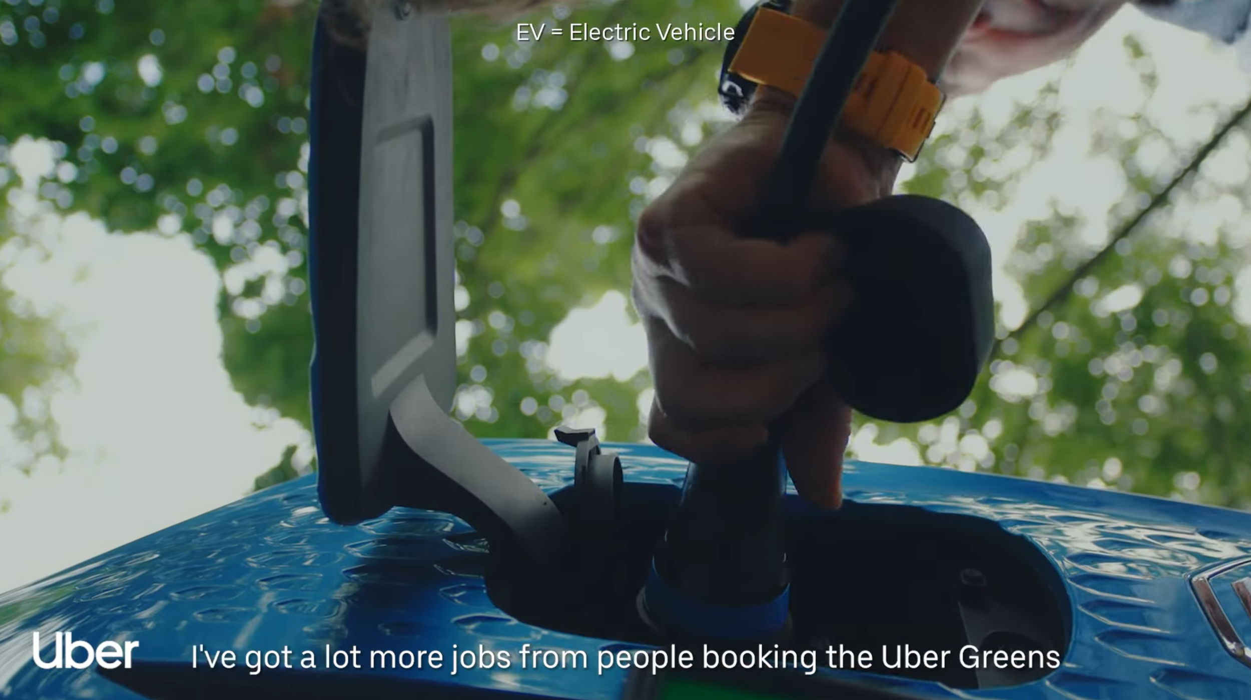 Close-up of a hand installing a charging cable into an electric vehicle's charging port, with trees and sky in the background.
