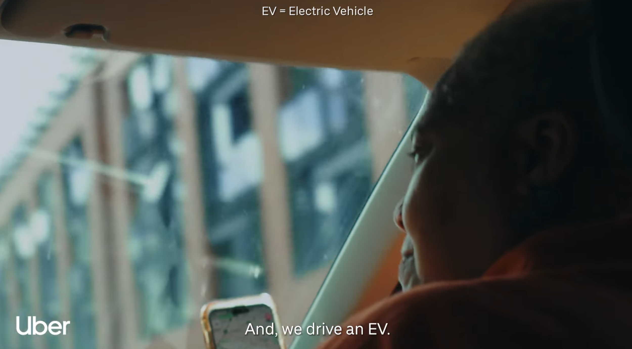Person sitting in the backseat of a vehicle, looking at a smartphone, with a blurred view of a construction site outside the window. Text on the image indicates the vehicle is an electric vehicle, with Uber branding.