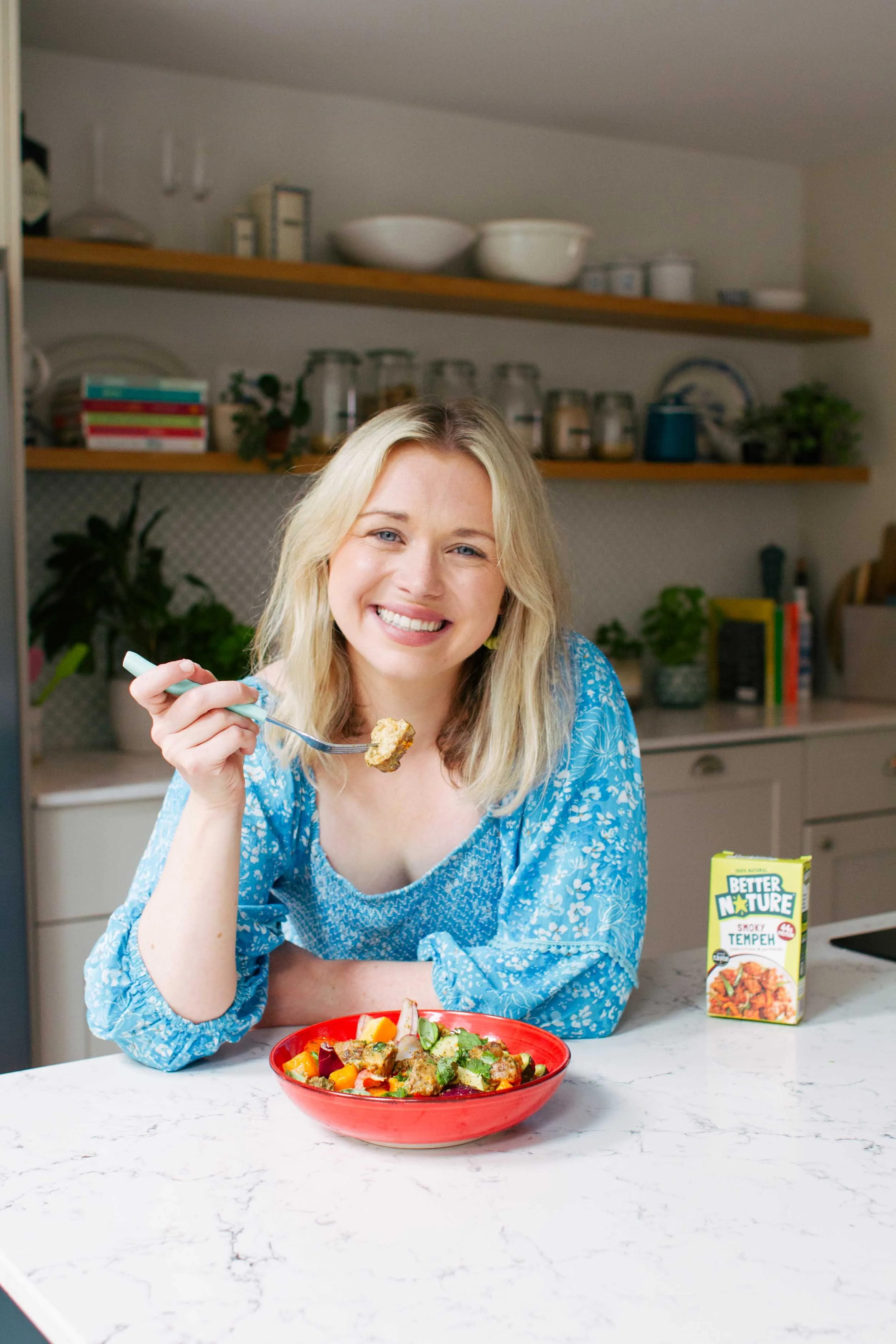 Food business owner in blue dress at kitchen counter with forkful of food and tempeh product, brand photography