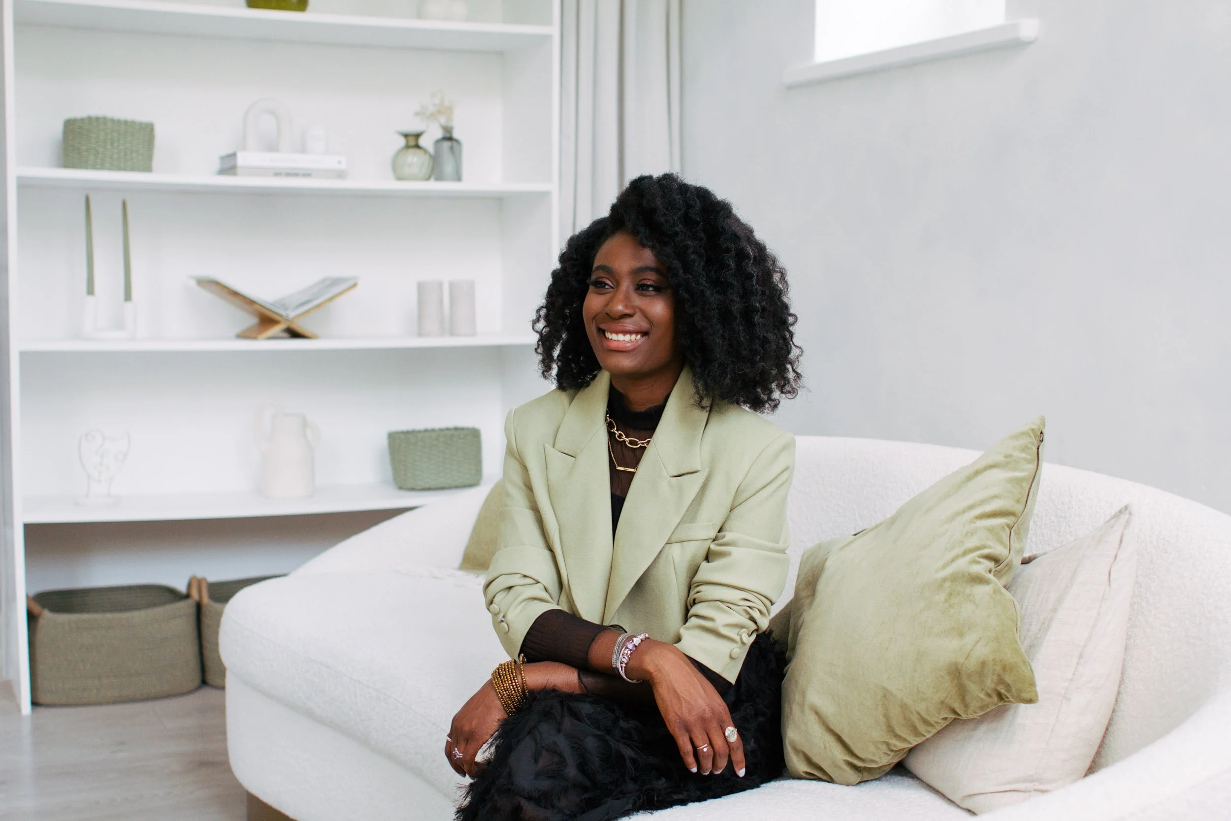 Woman in green blazer and black dress sitting on sofa with shelves behind her, brand photography