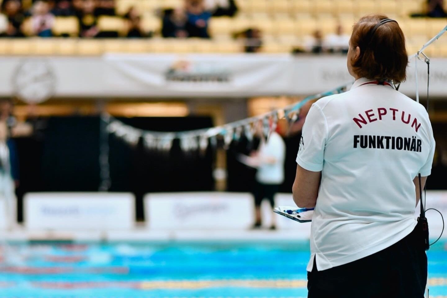 En kvinna i vit skjorta med texten 'NEPTUN FUNKTIONÄR' står vid en simbassäng och observerar ett simtävling.