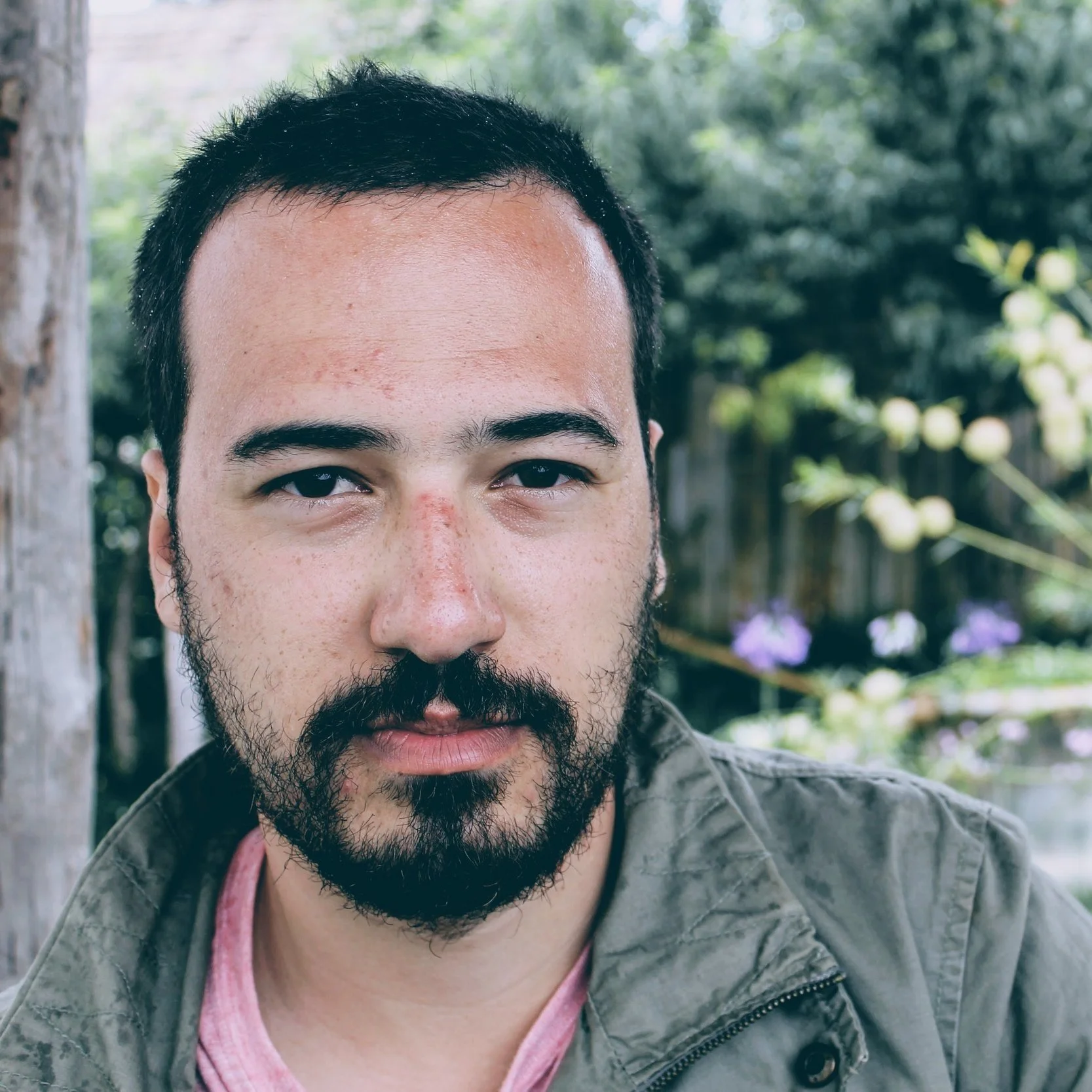 Close-up of a man with dark hair and a beard, wearing a green jacket, outdoors with blurred greenery and flowers in the background.