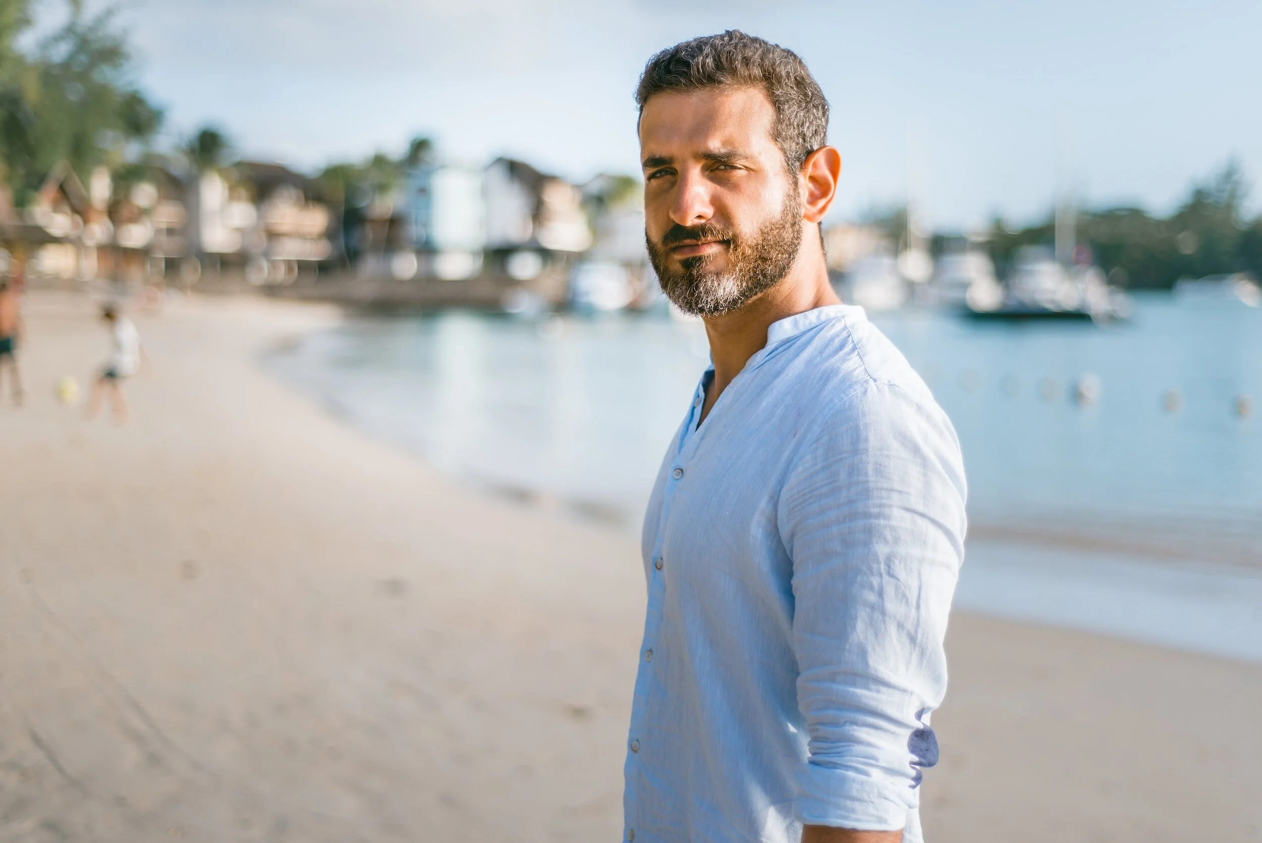 A man with a beard and short, dark hair wearing a light blue shirt standing on a sandy beach with boats and houses in the background.