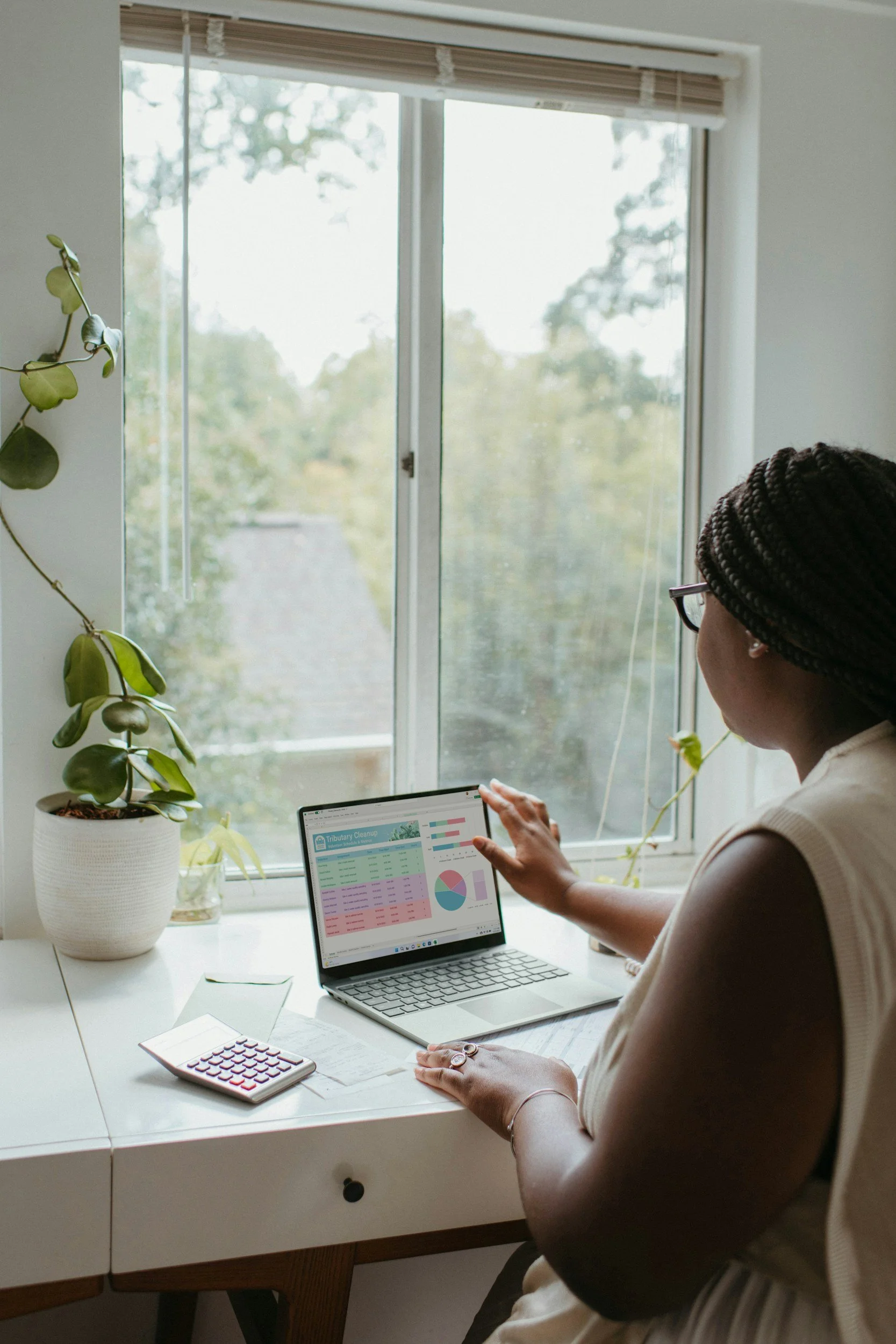 A woman sitting at a white desk near a window, working on a laptop displaying a spreadsheet and pie charts.
