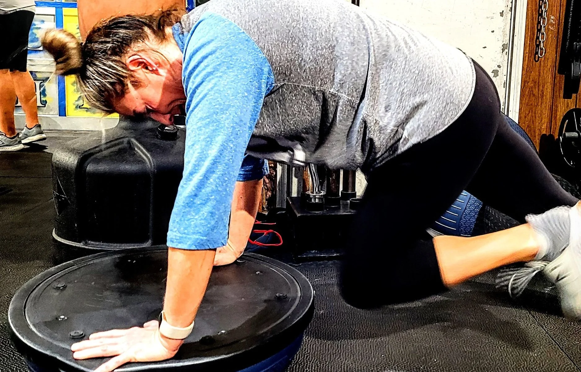 A woman using a vibration plate machine for exercise in a gym, kneeling on the floor with her hands on the machine's surface, wearing a gray and blue athletic outfit.