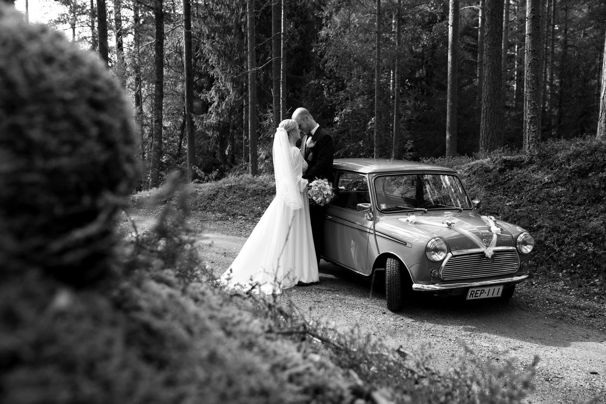 Häät kuva, pari tanssii läheisyydessä, vanha auto koristeltu kukilla, metsä taustalla.
