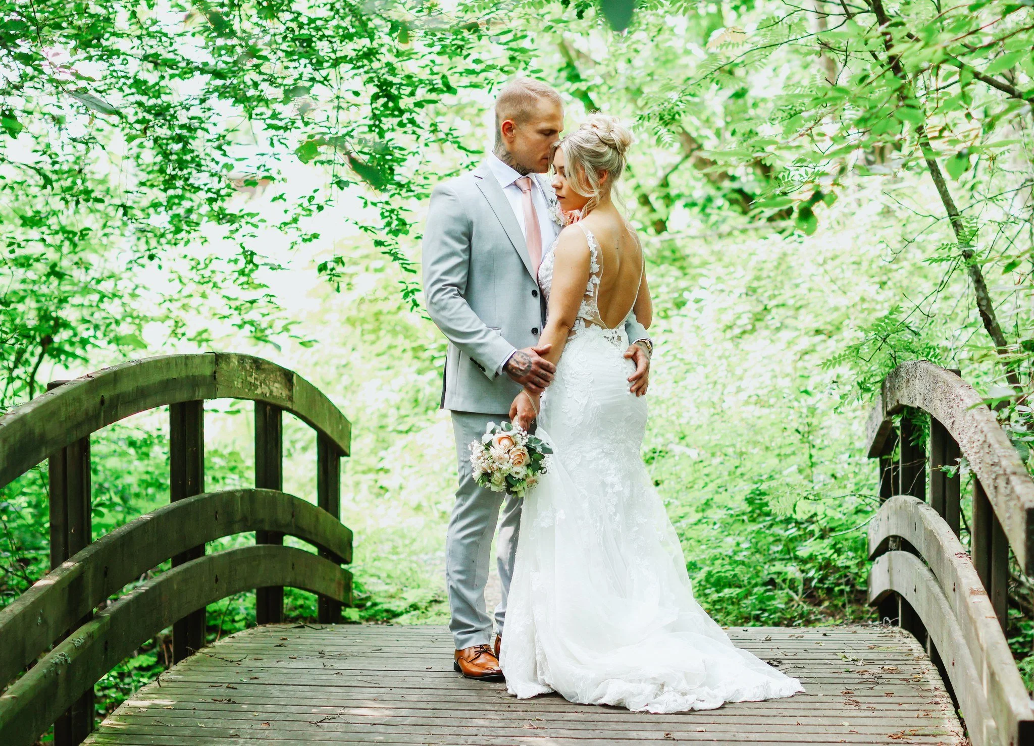 Hääpari seisoo pienellä puusillalla, ympärillään vihreää luontoa, nainen pukeutuneena valkoiseen hääpukuun ja mies harmaaseen pukuun, nainen pitää kukkakoristettua kimppua, he katsovat toisiinsa läheisesti.