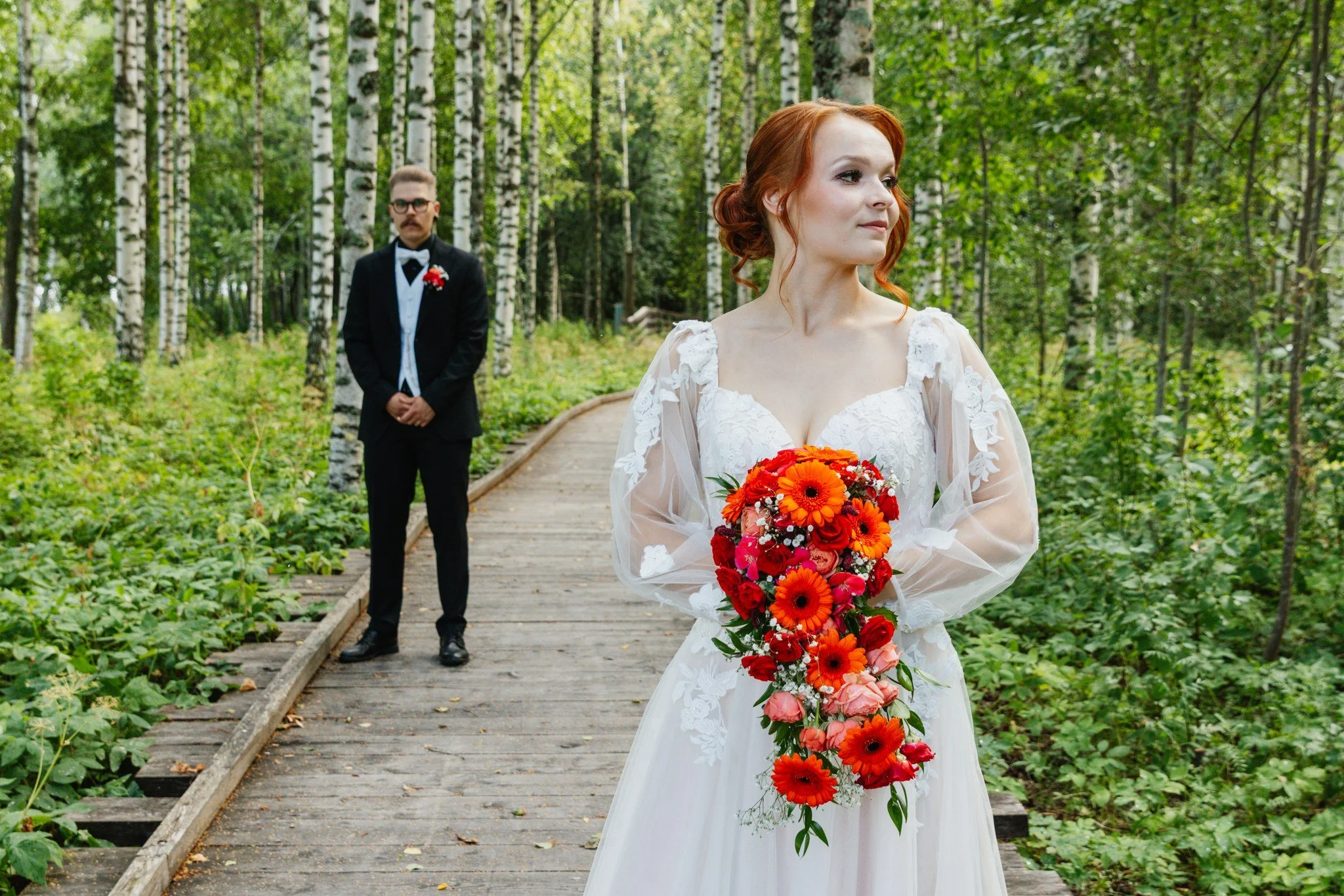 Häätikuva metsässä, morsian pitelee isoa kukkakoristetta, sulhanen seisoo taustalla.