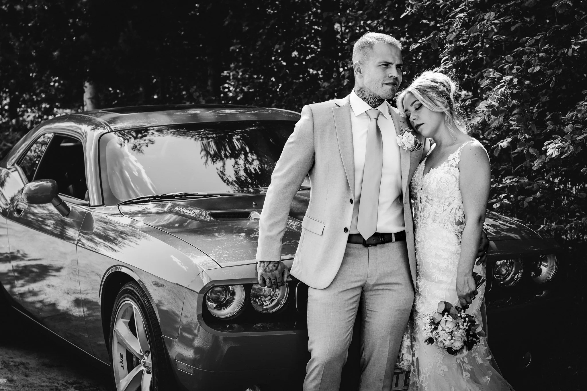 Häätunnelmallinen kuva, jossa sulhanen ja morsian seisovat toistensa vieressä auton ja puitten keskellä, molemmat hymyilevät ja pitävät kukkaseppelettä.