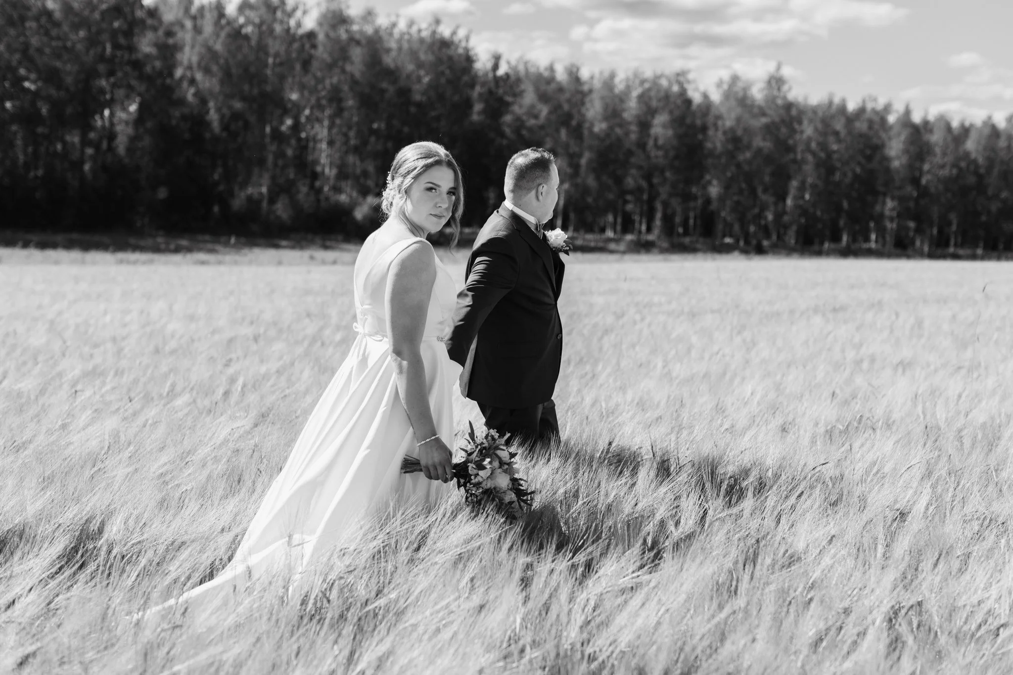 Hääpari kävelee pellolla, nainen pitää kukkakimppua ja miehellä on pikkutakki ja ruusuhattu, taustalla puita ja pilvinen taivas.