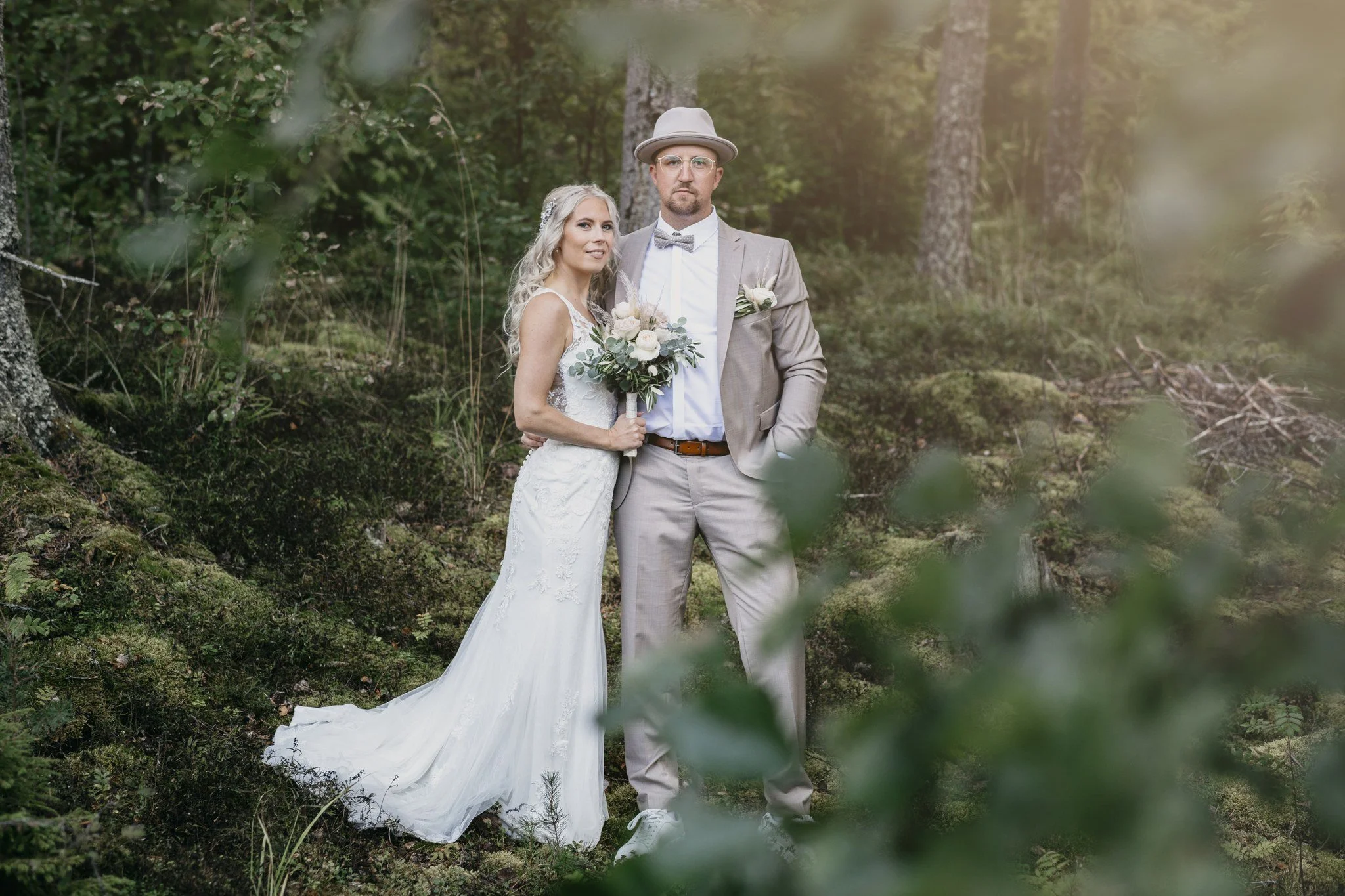 Hääpari seisoo metsikössä, nainen pitää kukkakimppua ja mies on pukuasussa hattuineen.