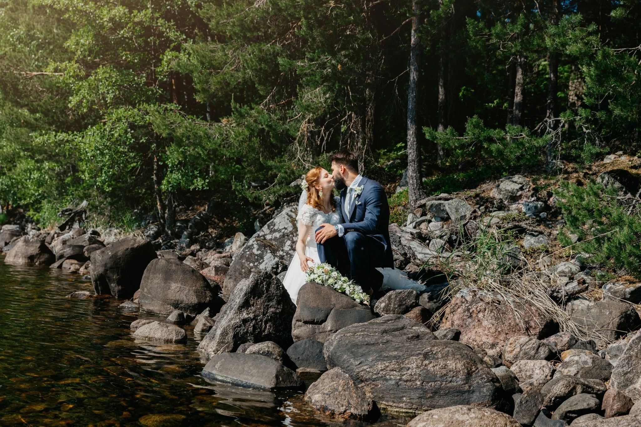 Hääpari istuu kallioilla järven rannalla metsä maisemassa.
