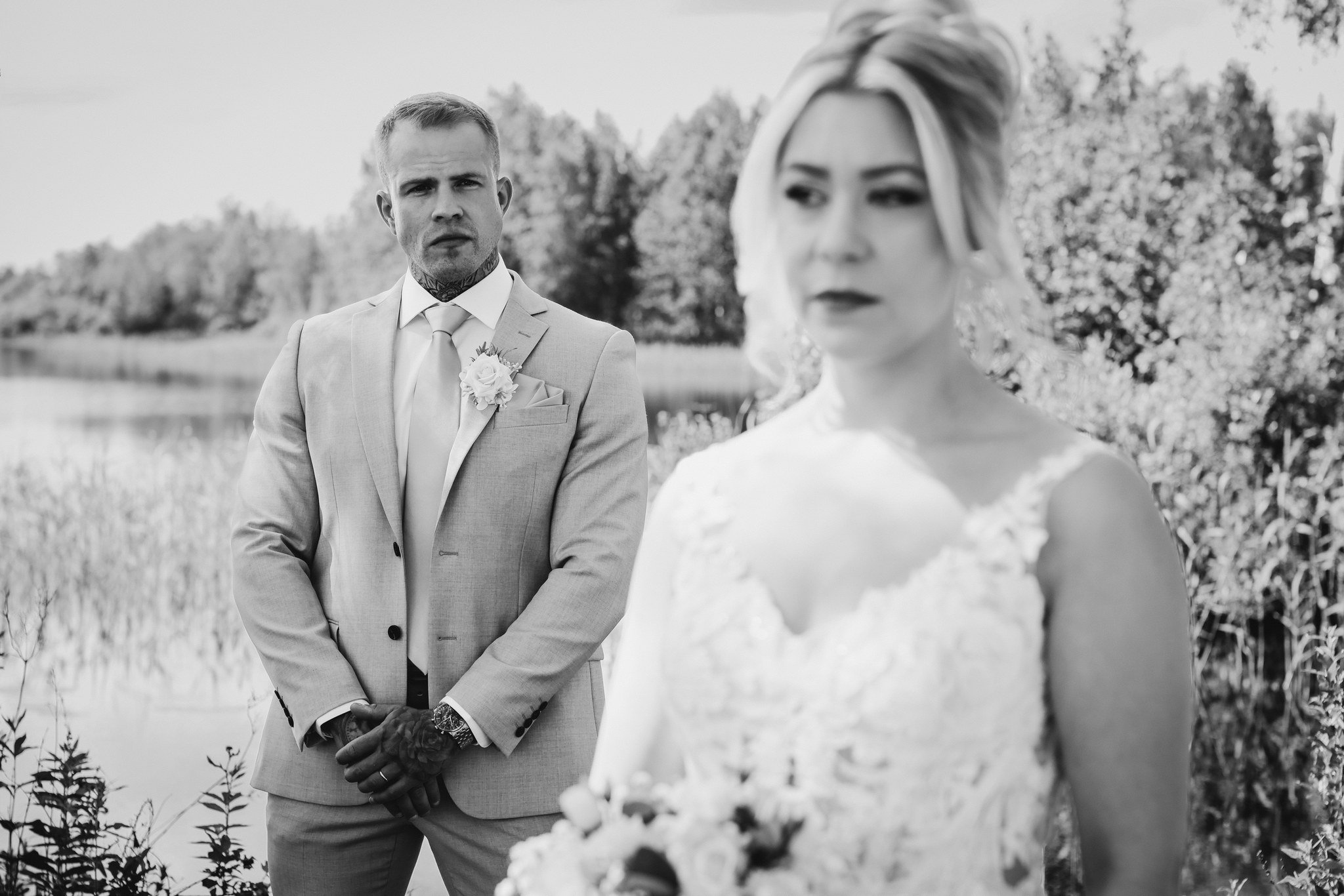 Valkoinen pukuinen mies katsoo naista, joka on epätarkka edessä. Taustalla on luonnonäkymä, joessa ja puissa.