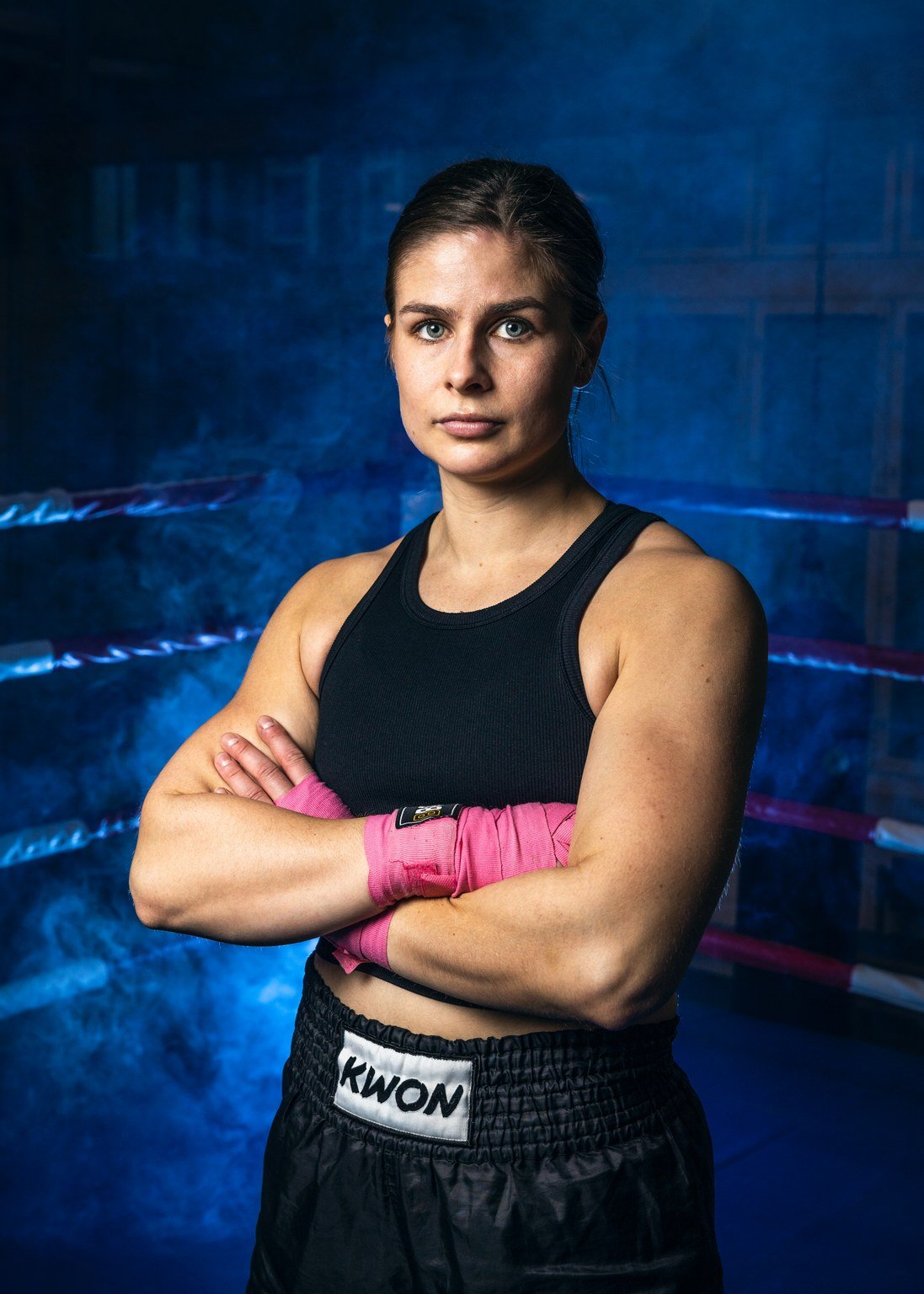 Naisten nyrkkeilijä poseeraa rintakehällä ristissä ja katsoo suoraan eteenpäin, taustalla sali ja musta seinä.