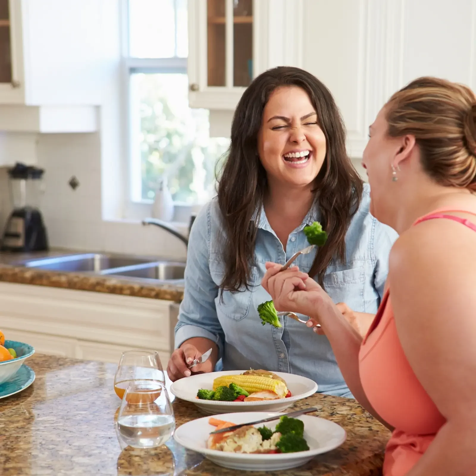 Two Women Smiling While Eating Together in a Kitchen