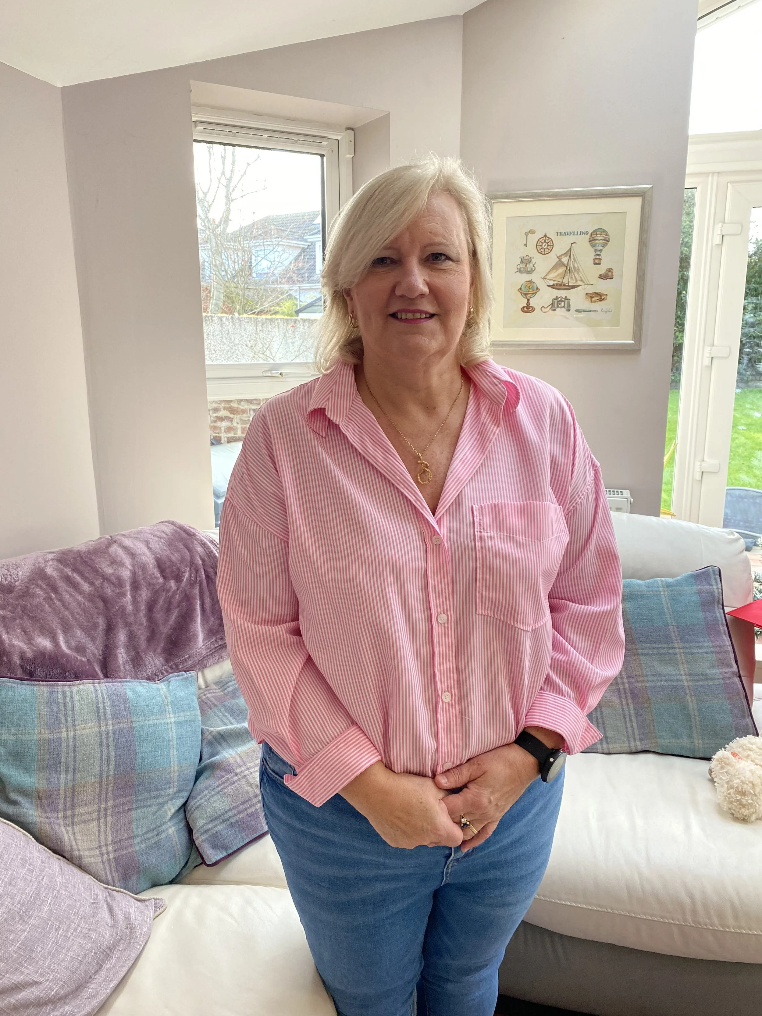 Smiling blonde woman with shoulder-length hair, wearing a pink shirt, standing in a bright room