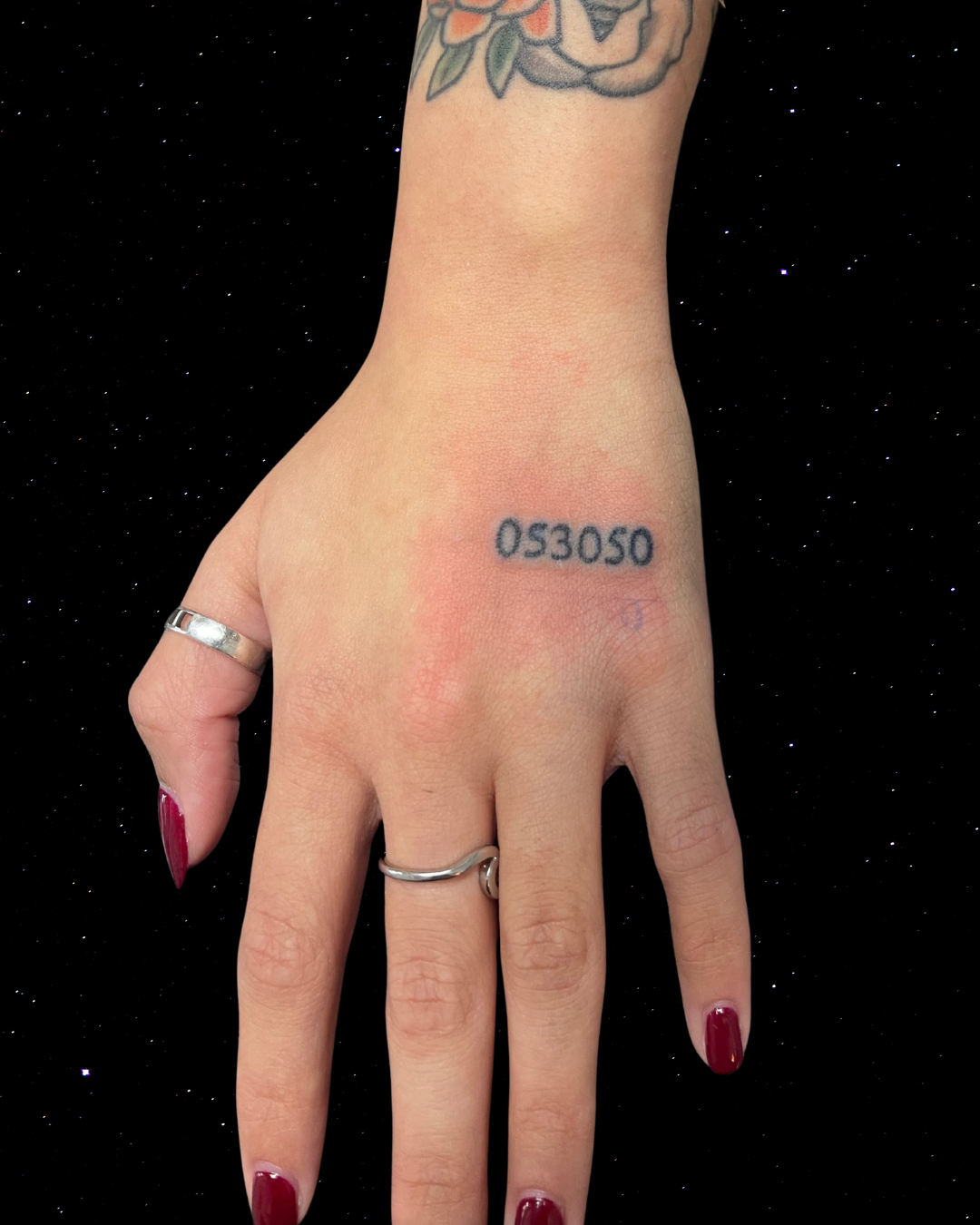 A person's hand with a hand poke tattoo of numbers. The hand has red painted nails and rings on two fingers. 