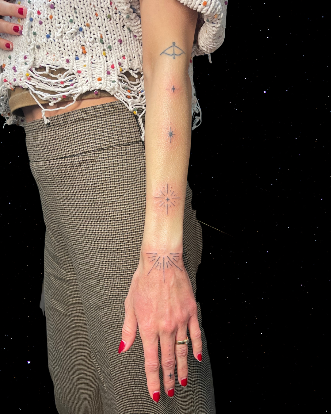 A person's arm and hand with five small, simple Hand Poked Tattoos along the arm and hand, wearing a frayed, colorful sweater and checkered pants