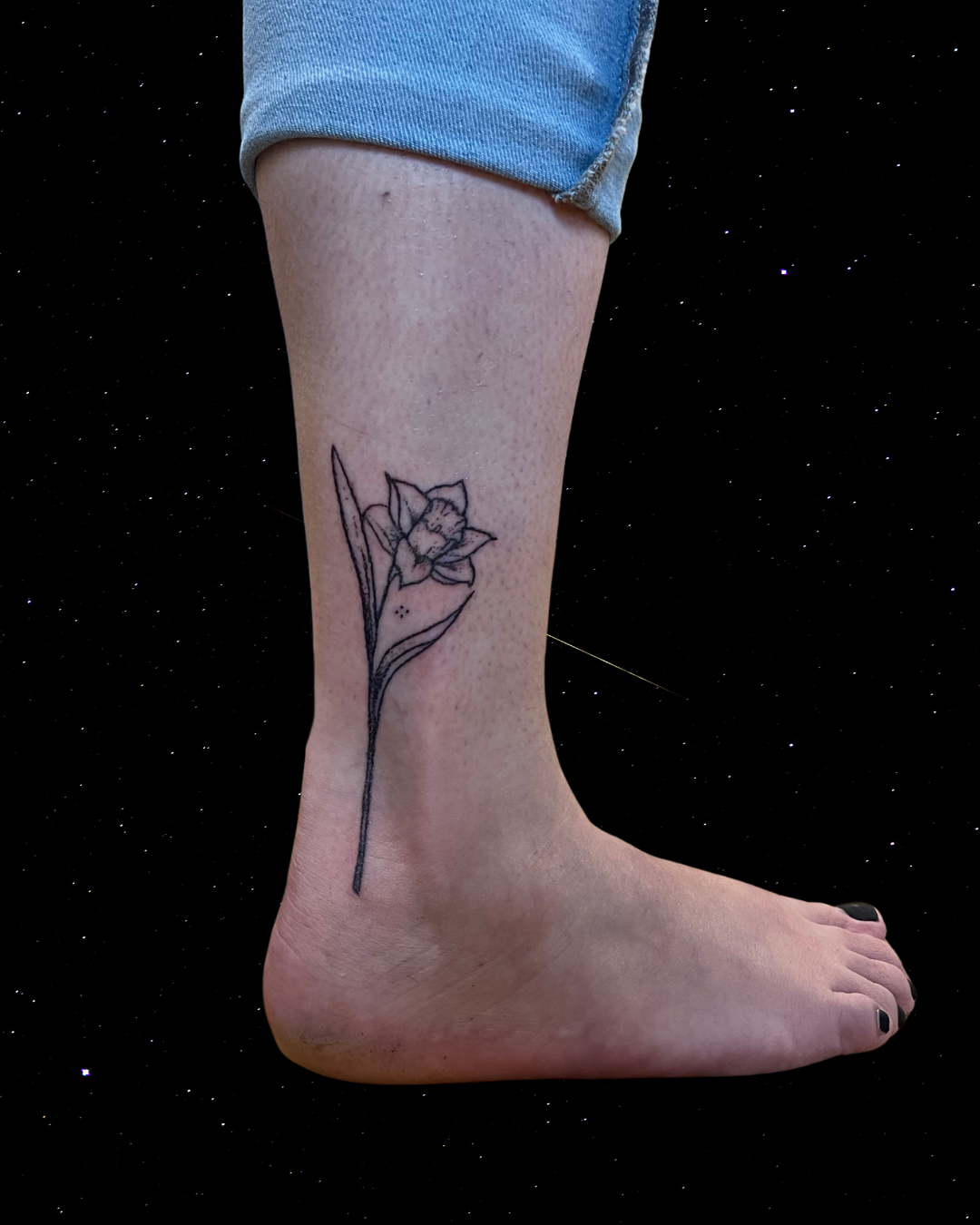 Hand Poked Tattoo of a daffodil flower on a person's lower leg and ankle, with a starry black background.