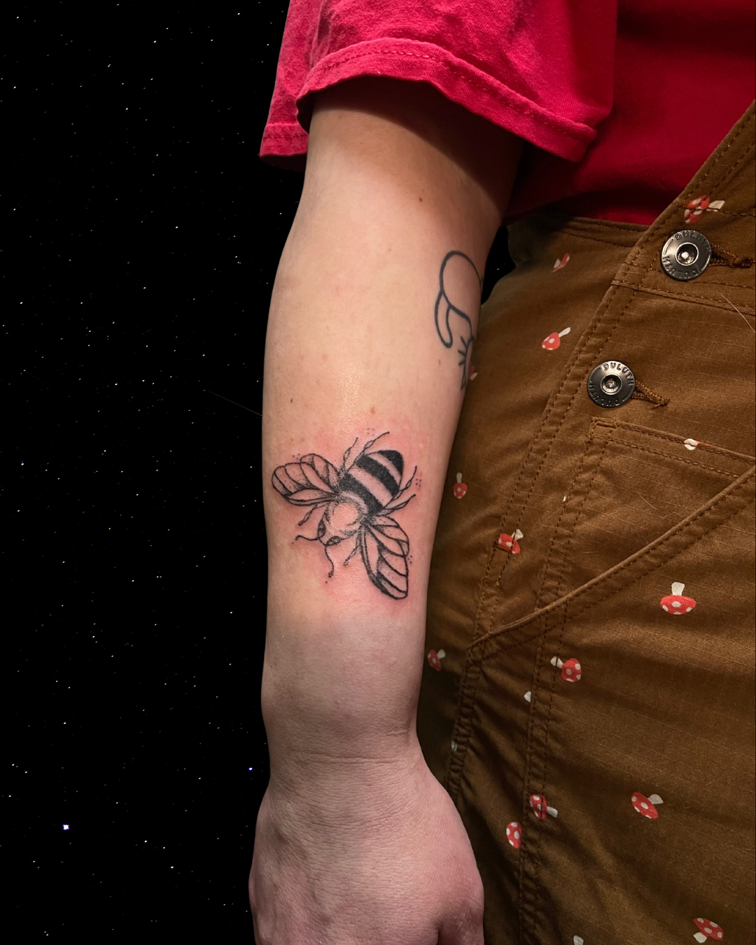 Hand Poked Tattoo of a bee on person's forearm, person wearing a pink shirt and brown pants with small mushroom patterns