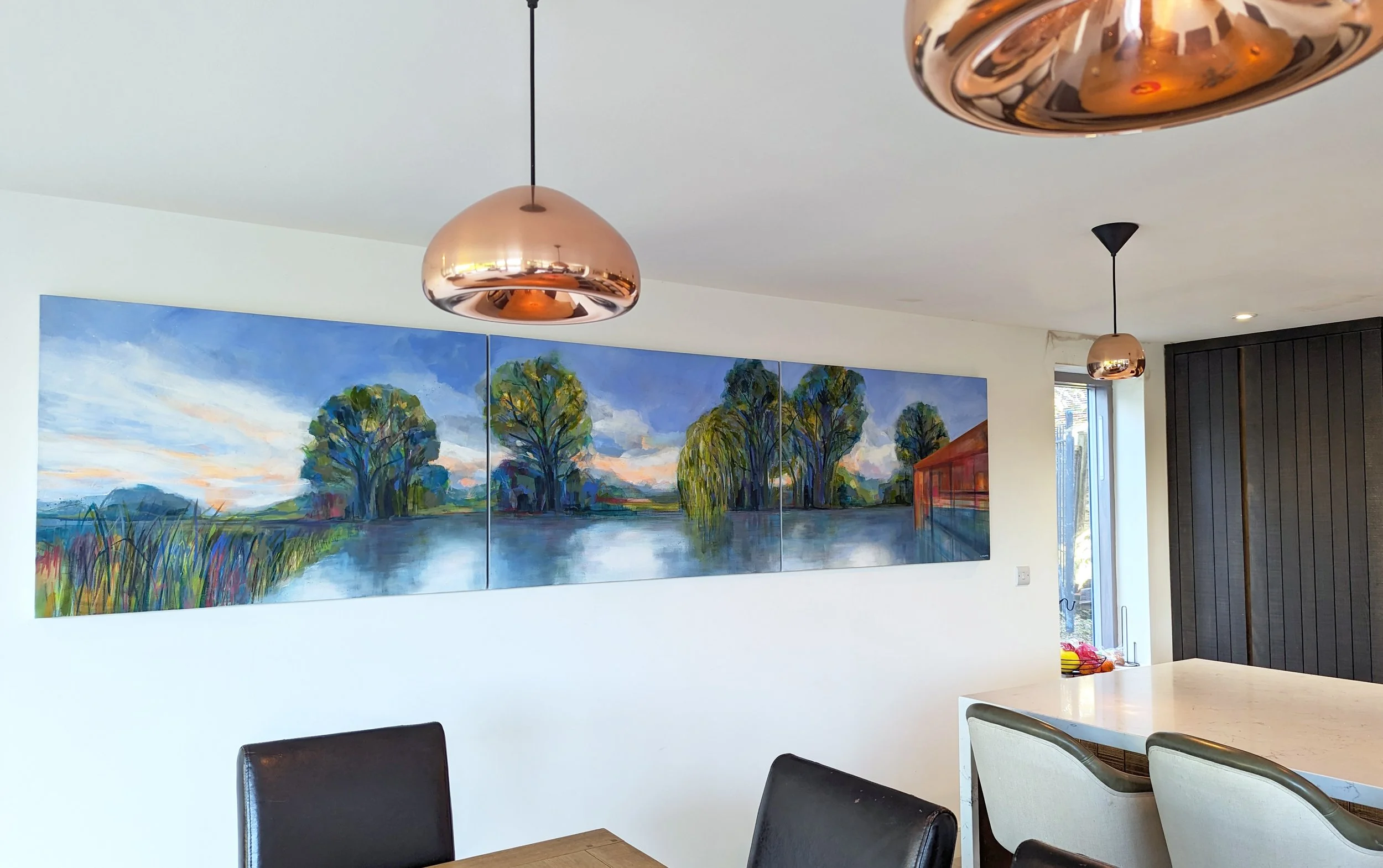 Interior of a dining area with a riverside landscape painting by artist Amy Wormald on the wall, copper pendant lights hanging from the ceiling, and a mixture of black and beige chairs around the table.