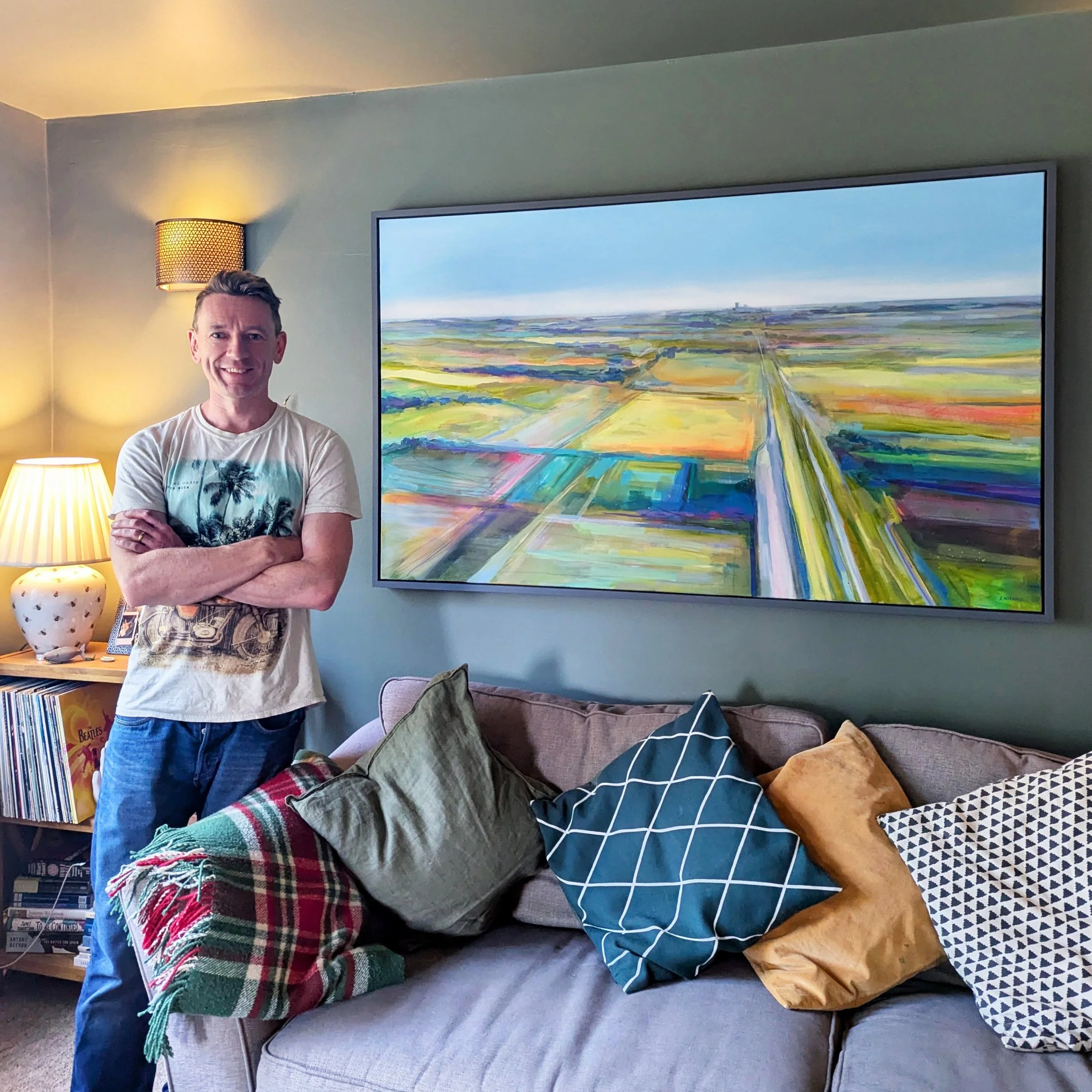 A smiling man with crossed arms standing next to a colourful abstract landscape painting by artist Amy Wormald in a living room with a sofa and various pillows.