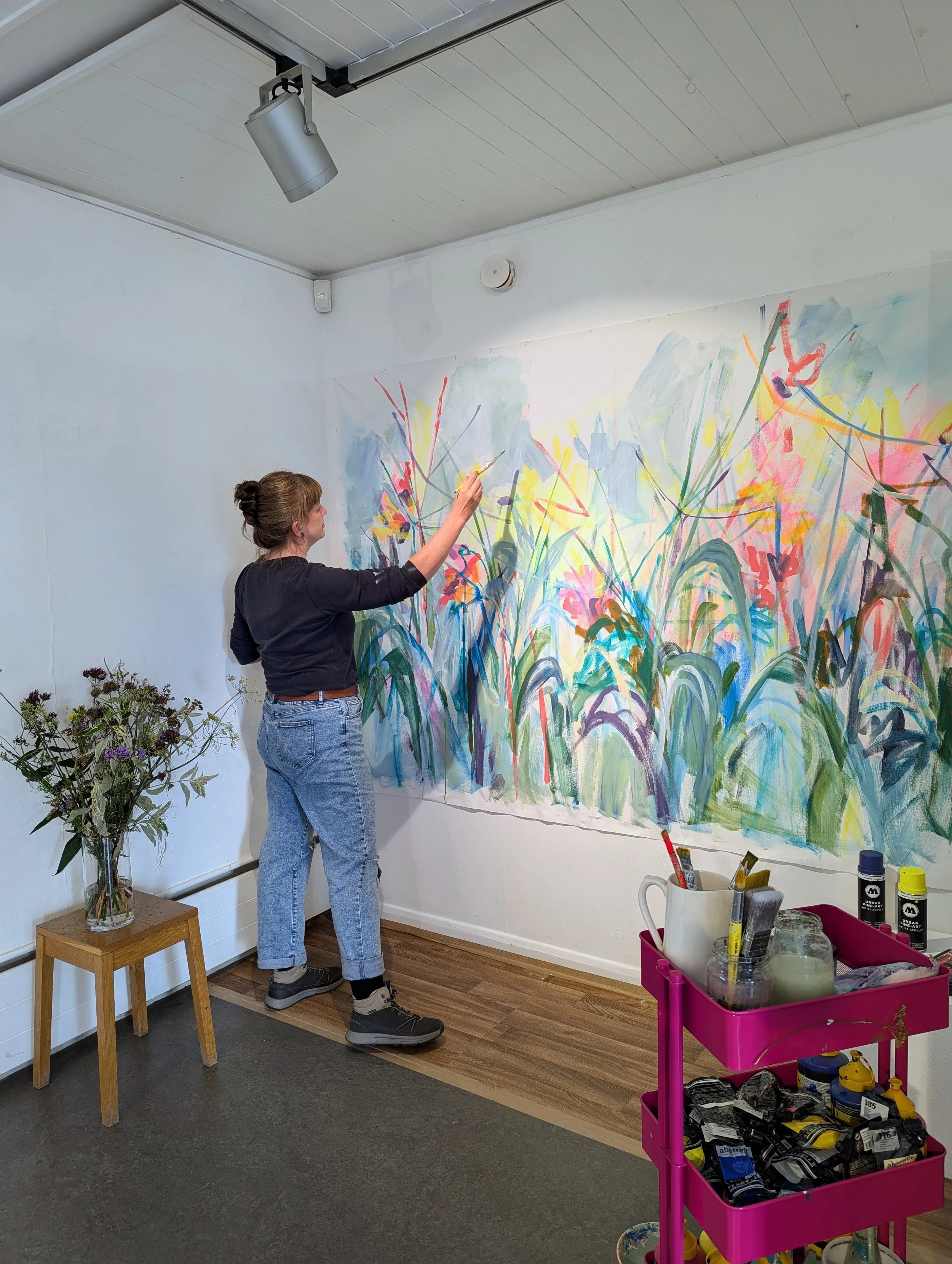 Artist Amy Wormald painting a colourful abstract mural on a wall in an art studio.