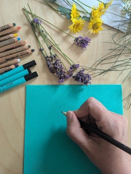 A person writing on a blue sheet of paper with a black pen, surrounded by colorful markers, dried lavender flowers, and yellow and purple flowers on a wooden surface.