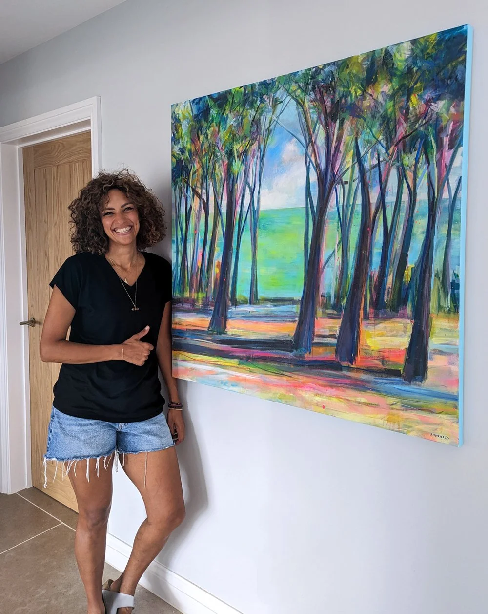 A woman with curly hair, wearing a black T-shirt and denim shorts, standing next to a colourful landscape painting by artist Amy Wormald on a white wall, in a room with a wooden door.