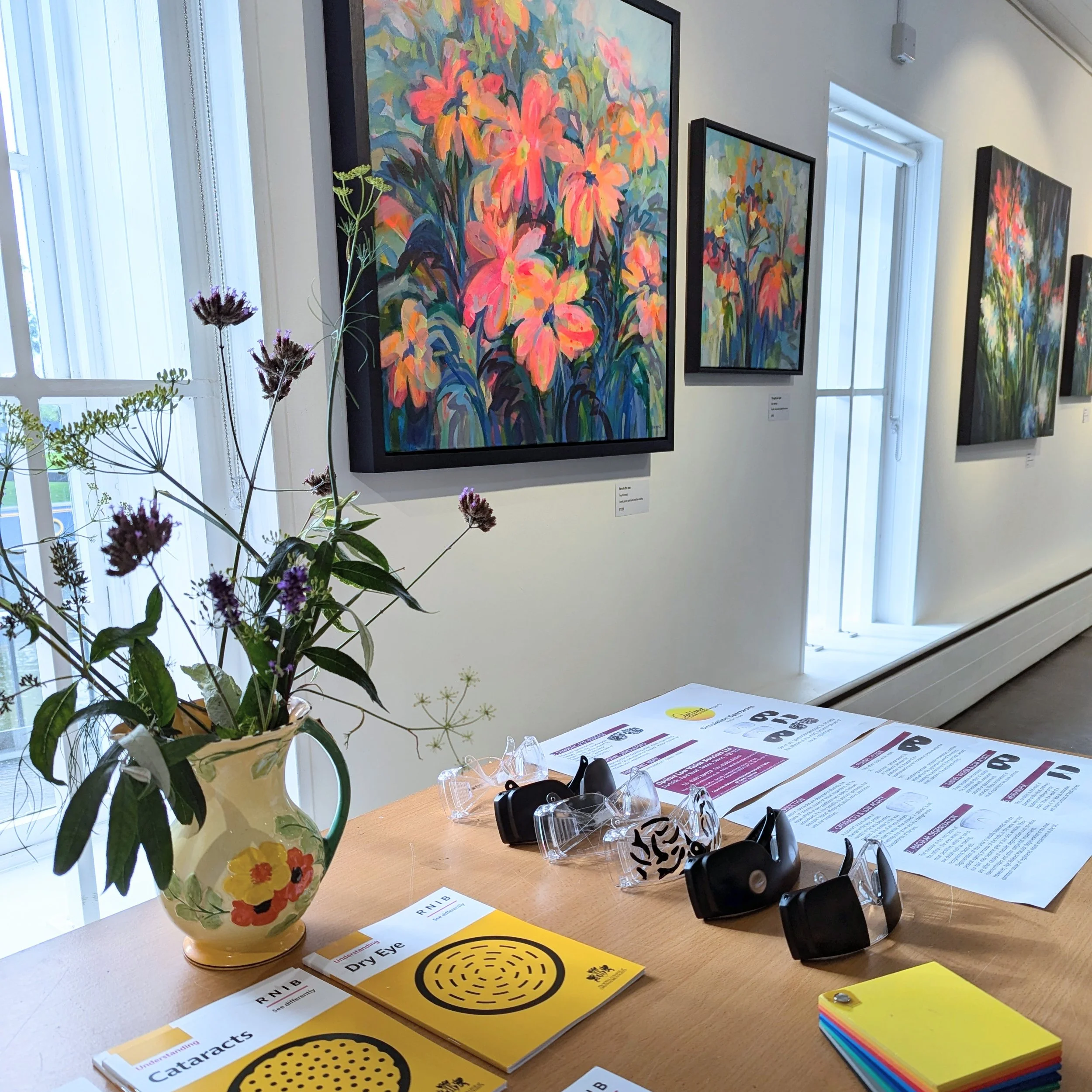 An art gallery with colorful floral paintings on the wall. A table with pamphlets, glasses, and keychains, and a vase with wildflowers near window.
