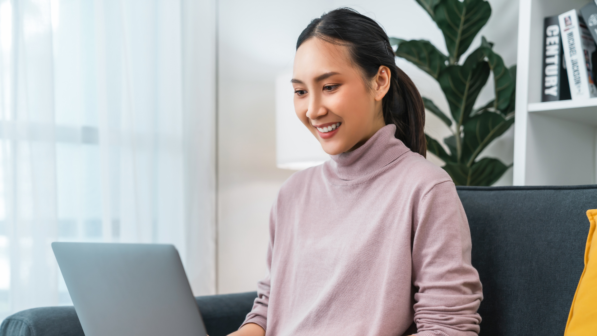 A young woman with dark hair in a ponytail, wearing a pink turtleneck, smiling while working on a laptop in a bright room with a large plant and a white bookshelf in the background.