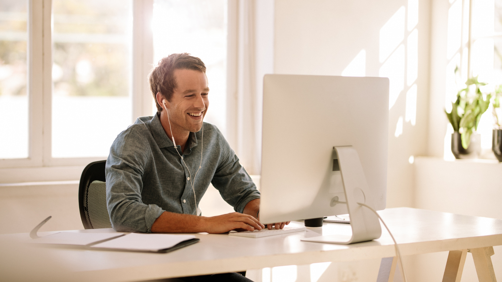 A man sitting at a white desk in front of a computer, smiling, wearing earbuds, with sunlight streaming through large windows in the background.