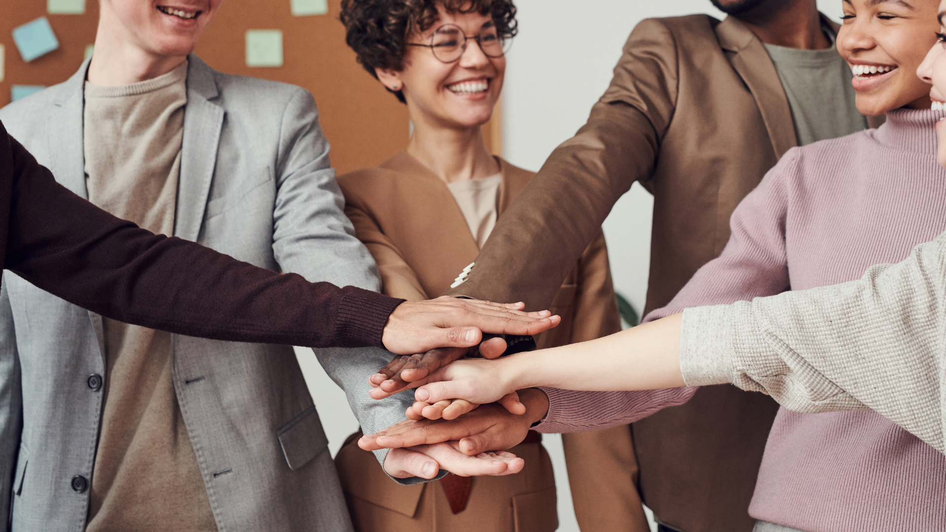 Group of diverse people placing their hands together in a team gesture, smiling, in an office setting.