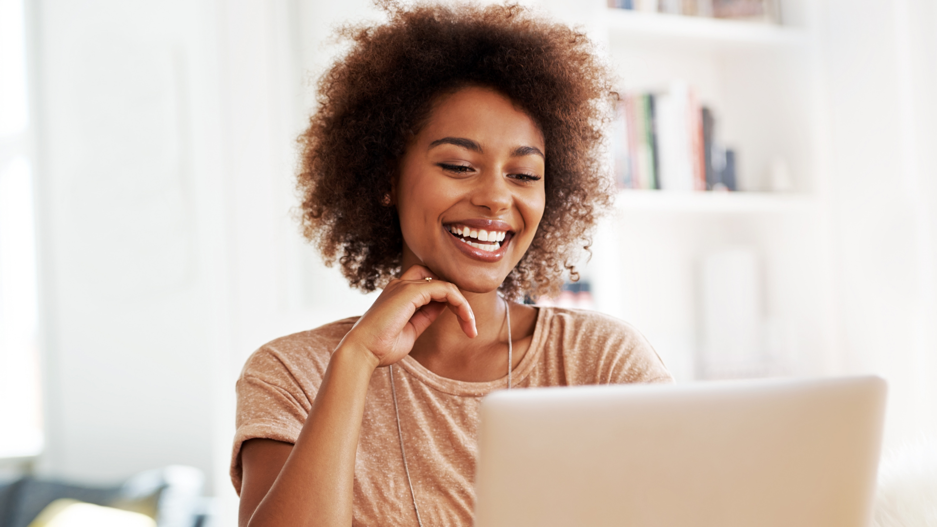 A woman with curly hair smiling and sitting in front of a laptop, possibly in a home or office setting.