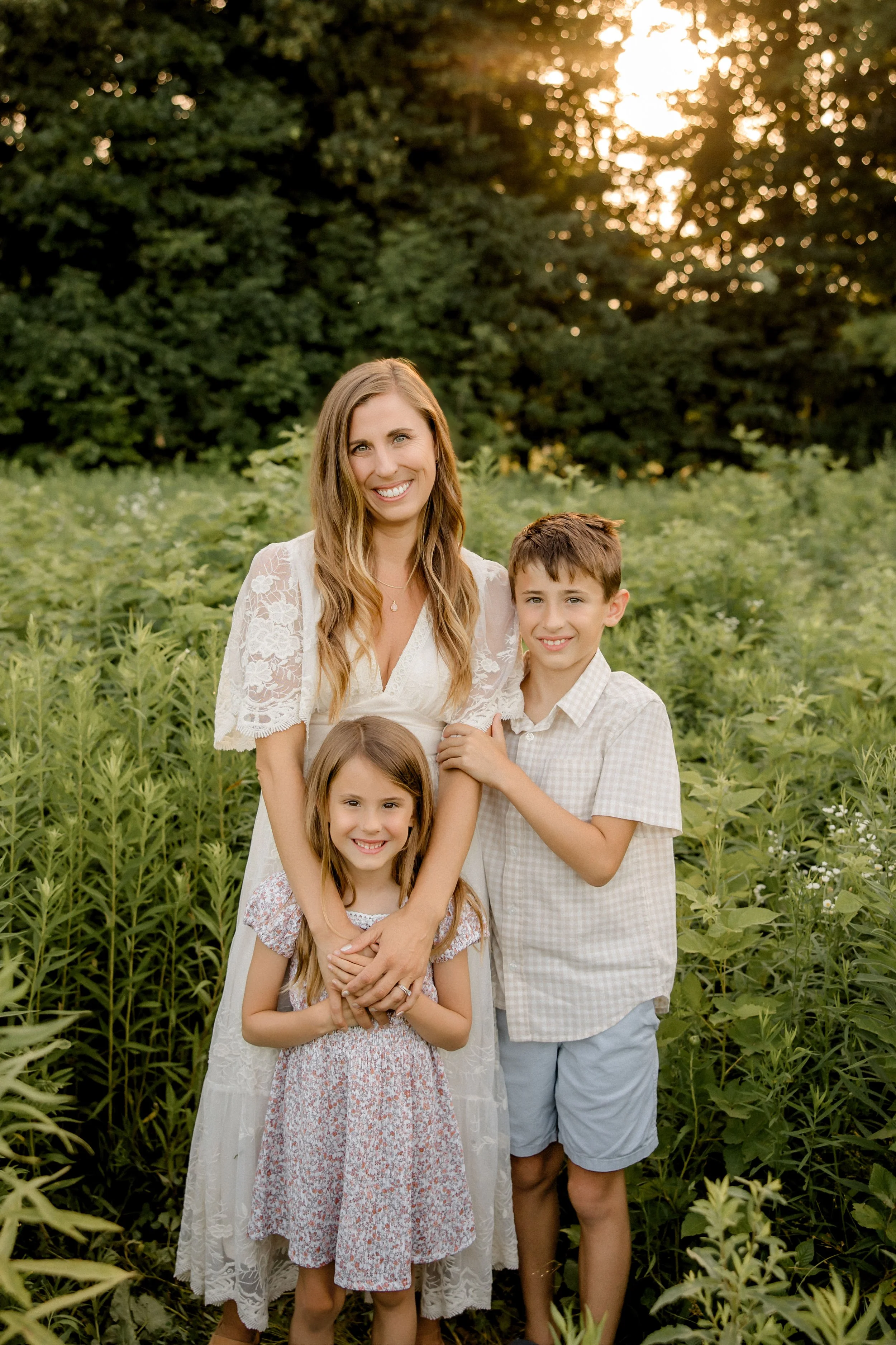 A woman with two children standing in a lush green field during sunset, smiling at the camera.