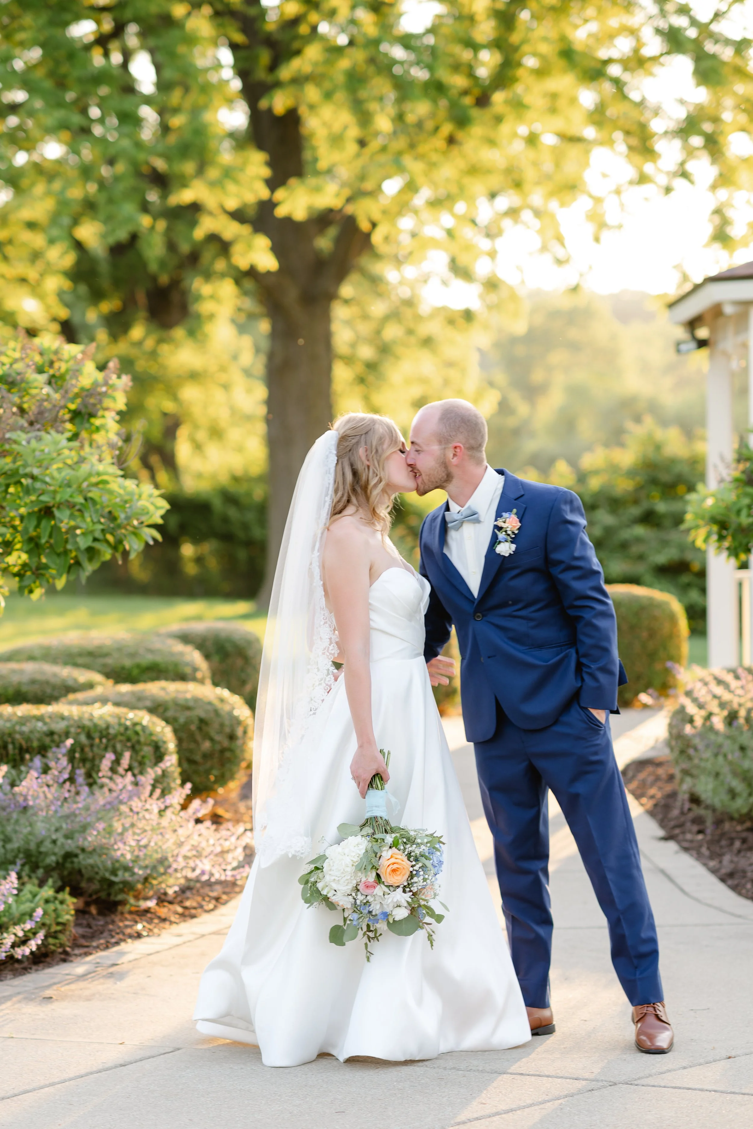 A bride and groom kiss outdoors in a garden setting, with lush green trees and blooming bushes in the background. The bride wears a white wedding gown, holds a bouquet, and the groom is dressed in a blue suit with a light gray bow tie.