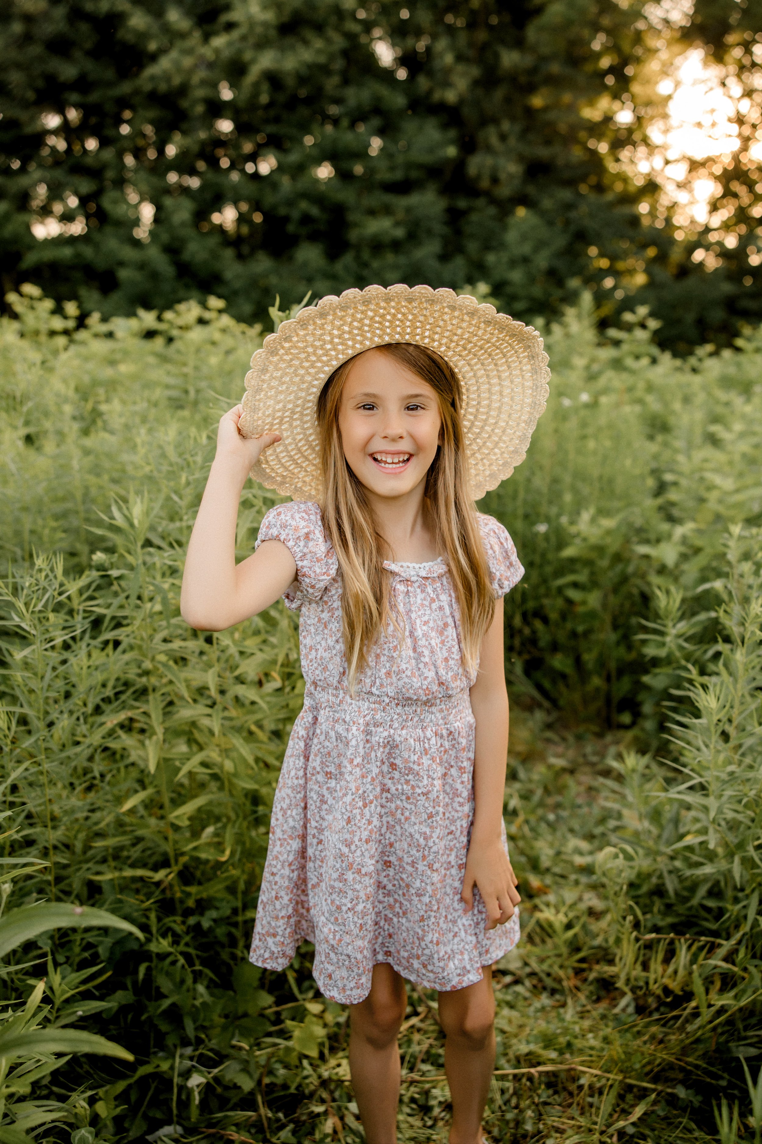 A young girl in a floral dress, smiling and holding a large straw hat, standing outdoors in a green field during sunset.